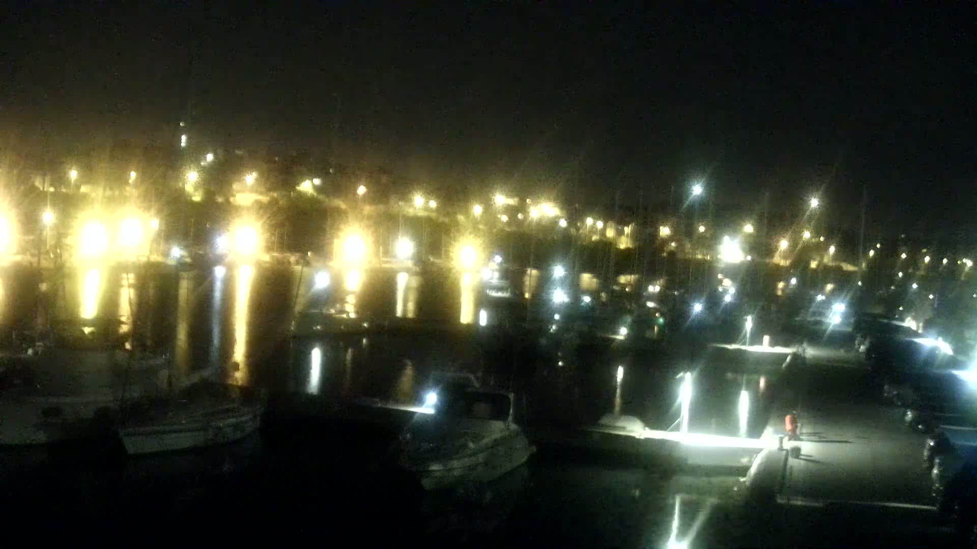 Many boats are docked at a marina at night, illuminated by lights reflecting on the water.
