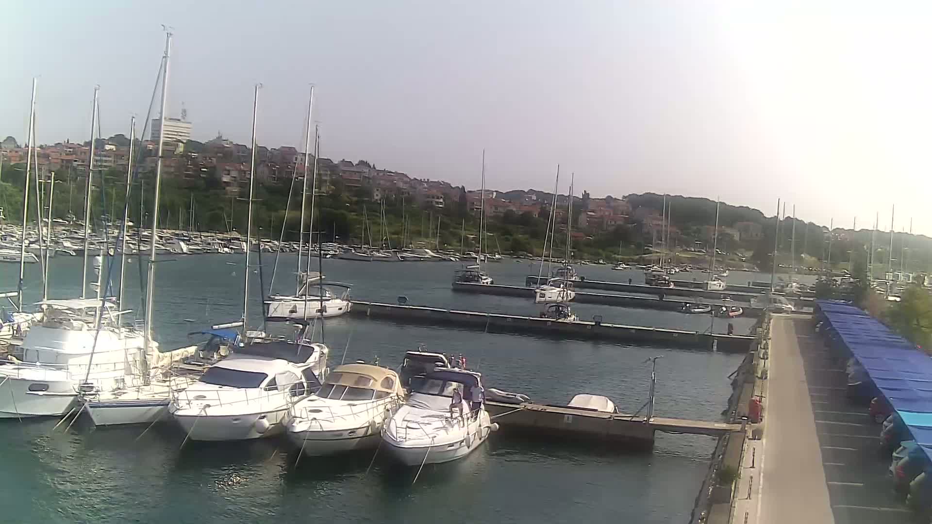 A marina filled with sailboats and motorboats on a calm, sunny day, with a town visible in the background.