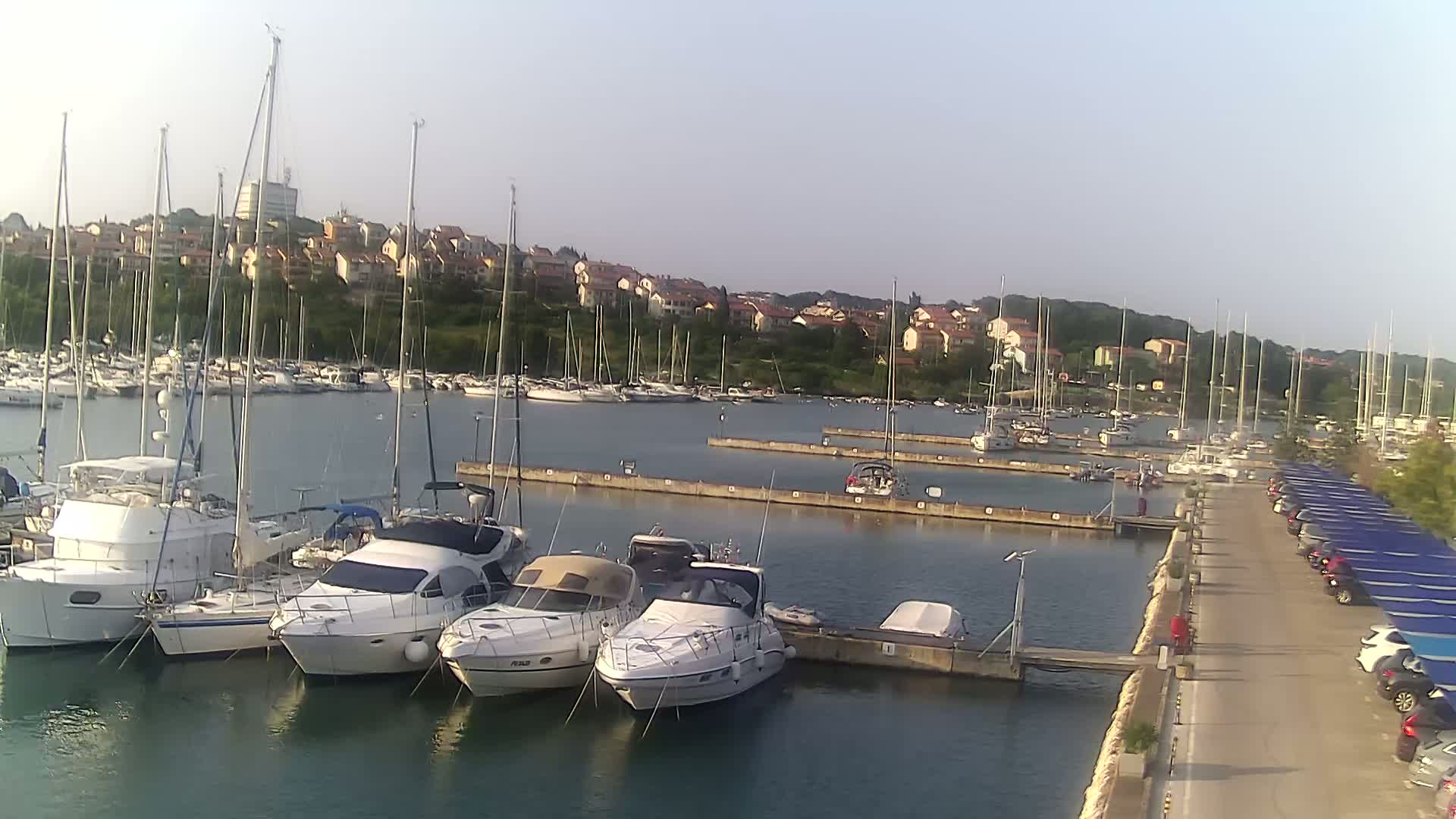 A marina filled with sailboats and motorboats on a calm, sunny day, with a residential area visible in the background.