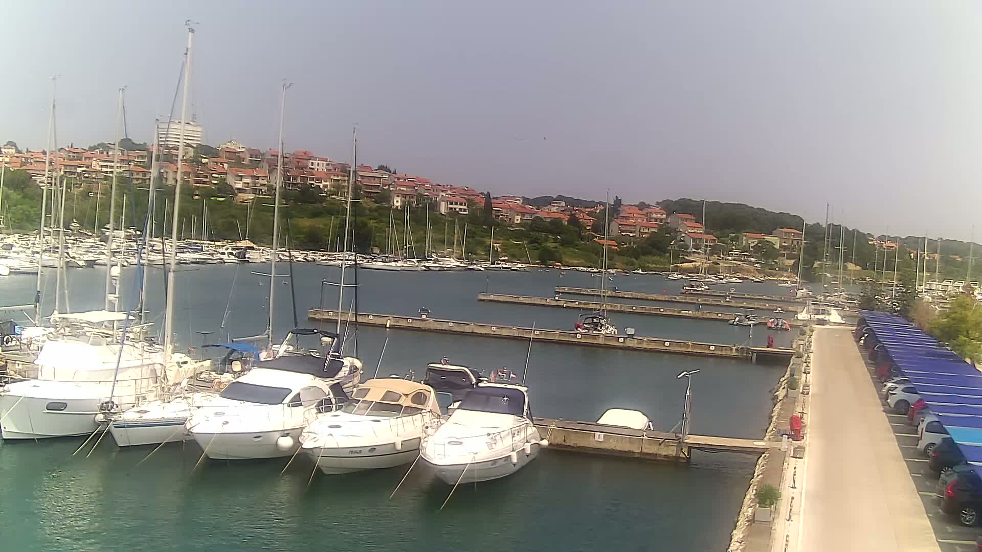 A marina filled with sailboats and motorboats is situated on calm water under a clear, sunny sky, next to a paved area with parked cars under blue canopies.
