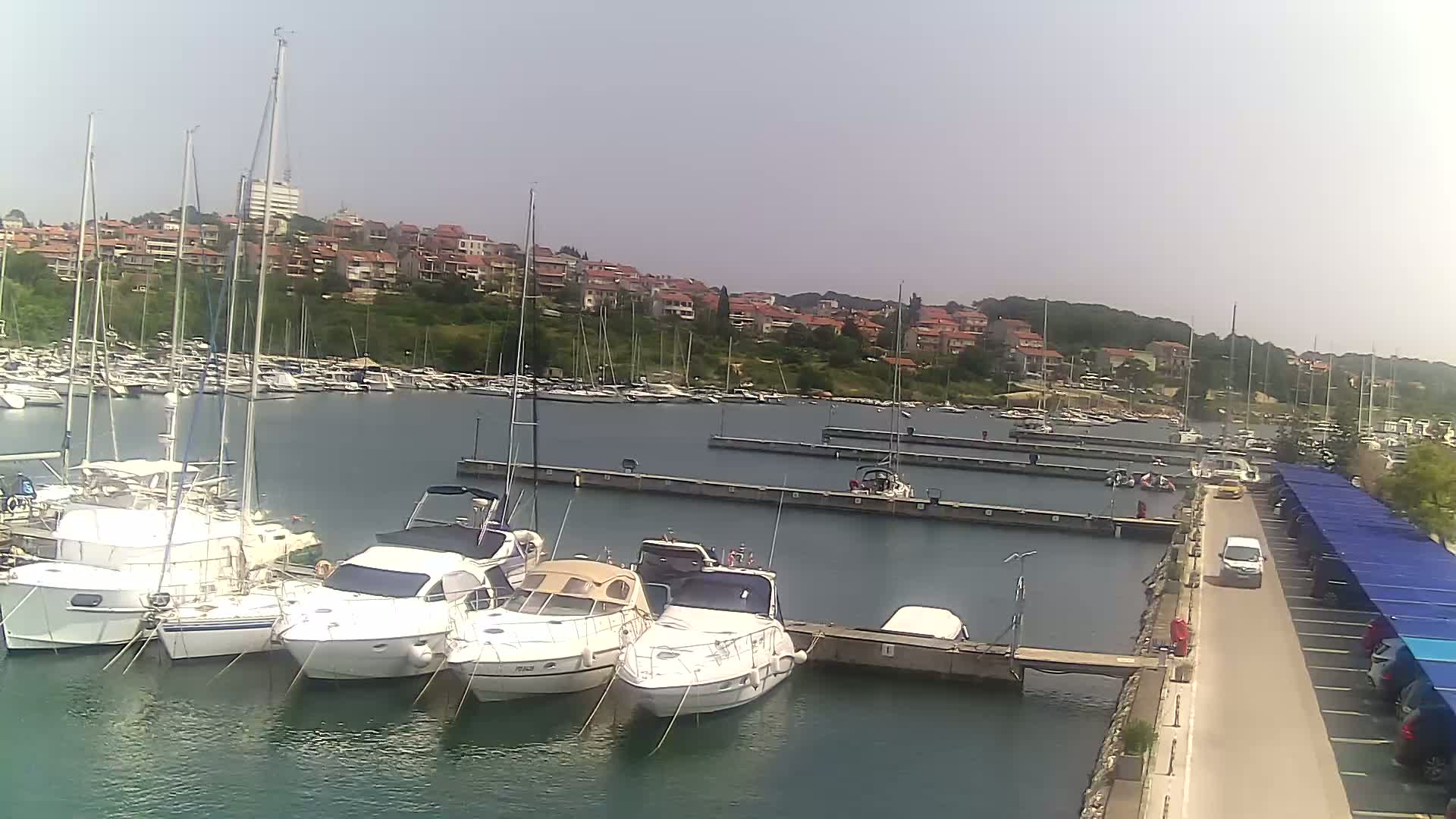 A marina filled with sailboats and motorboats on a calm, sunny day, with a residential area visible in the background.