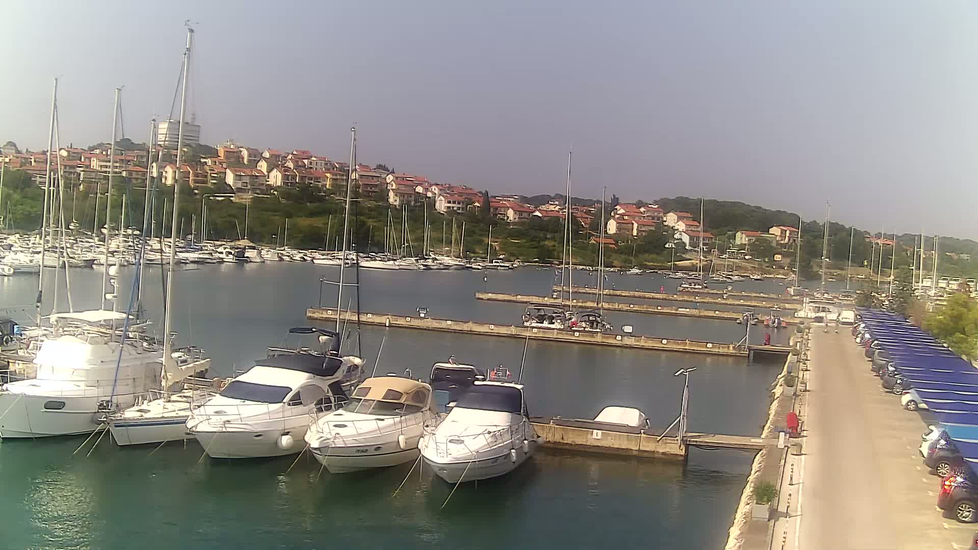A marina filled with numerous boats, some docked and others at anchor, sits in calm water under a clear, sunny sky, with a hillside of houses visible in the background.