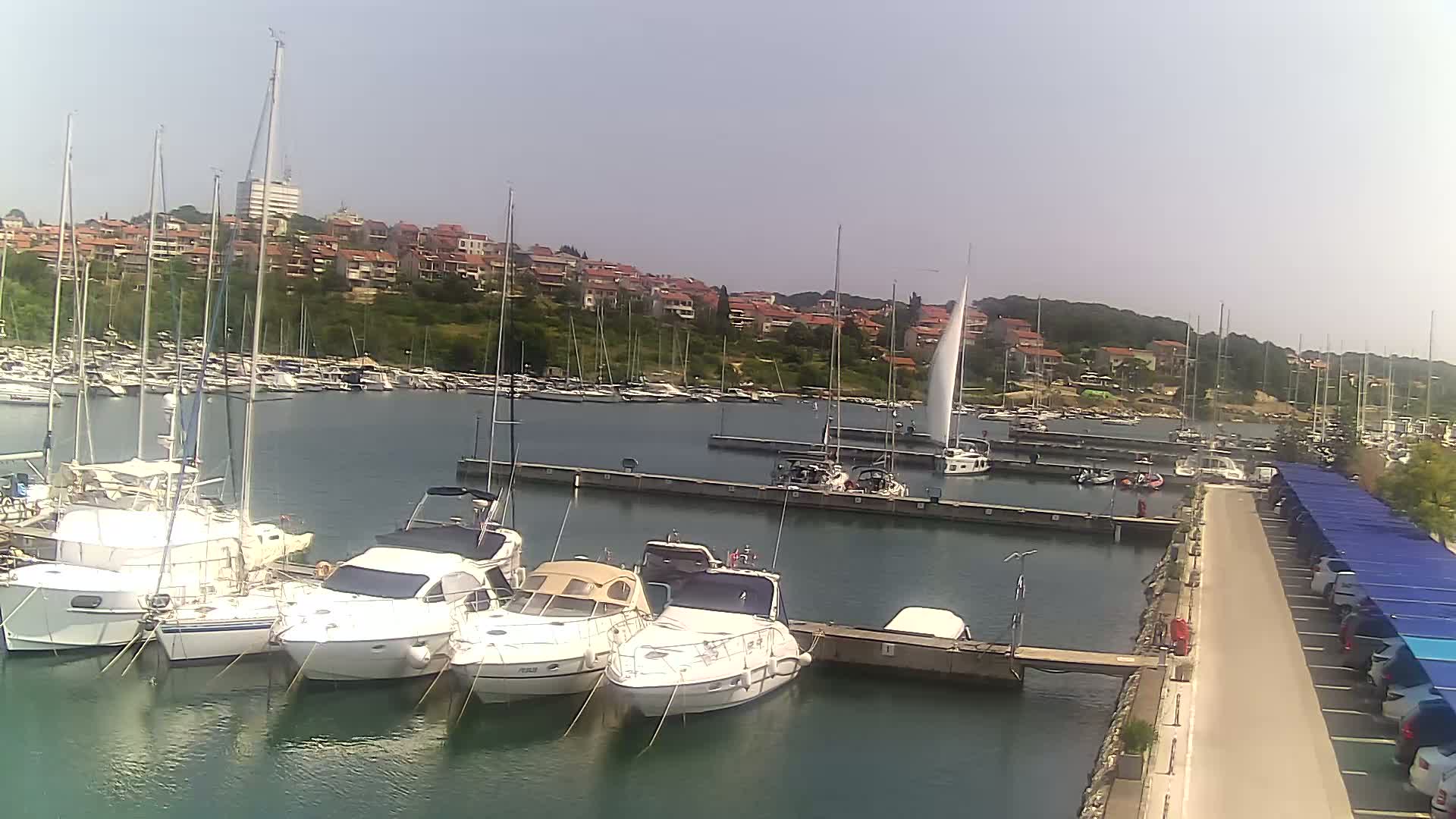 A marina filled with sailboats and motorboats is situated beside a paved walkway with covered parking under a sunny sky.