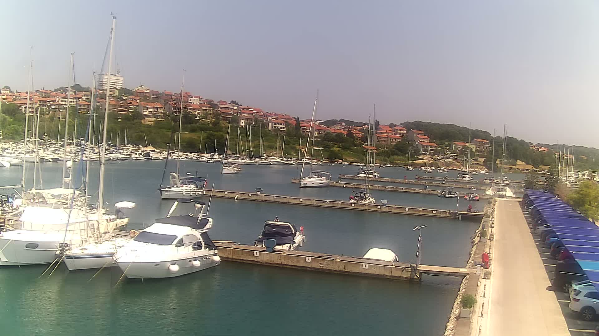 A marina filled with sailboats and motorboats on a sunny day, situated near a hillside town.