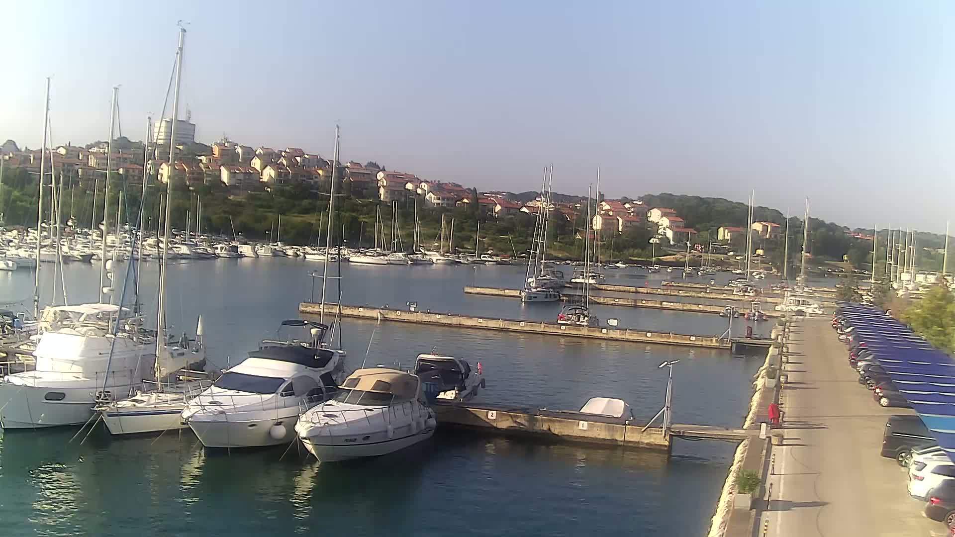 A marina filled with sailboats and motorboats is situated near a town of low-rise buildings on a sunny day.