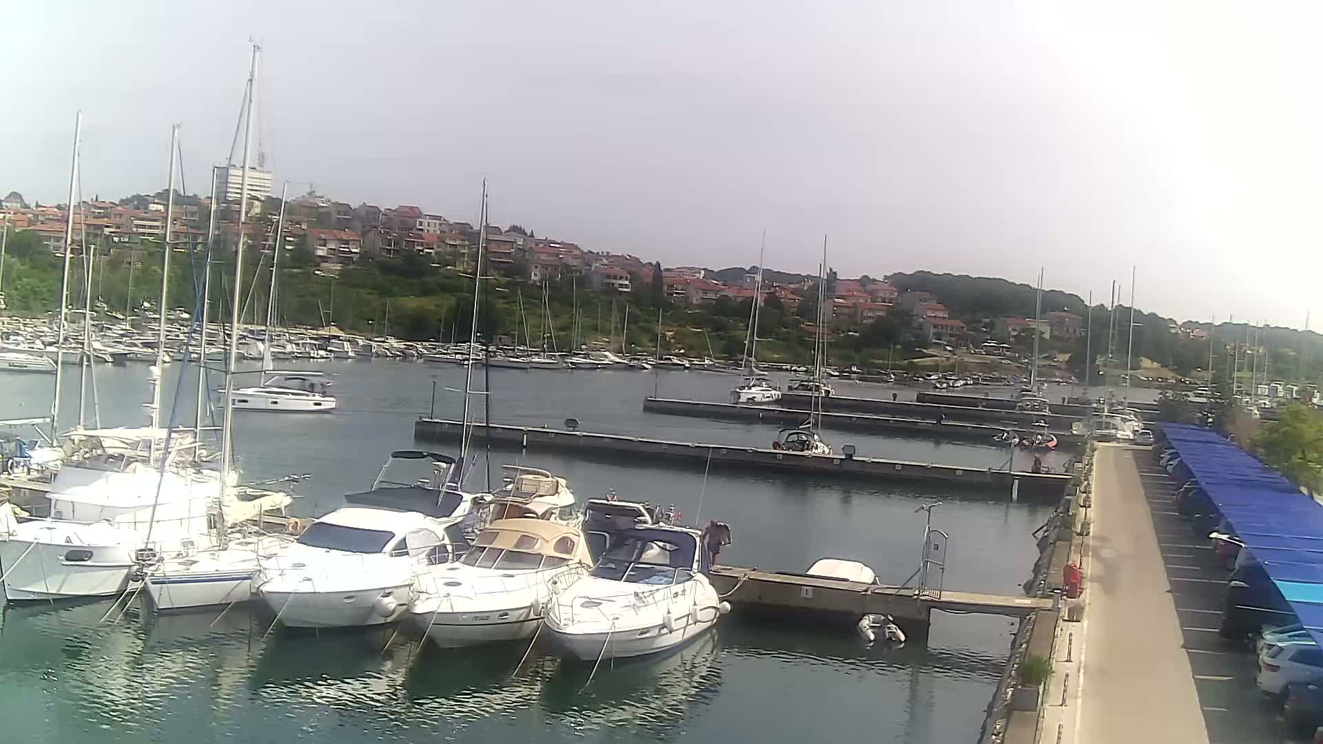 A marina filled with sailboats and motorboats is situated near a town under a mostly clear sky.