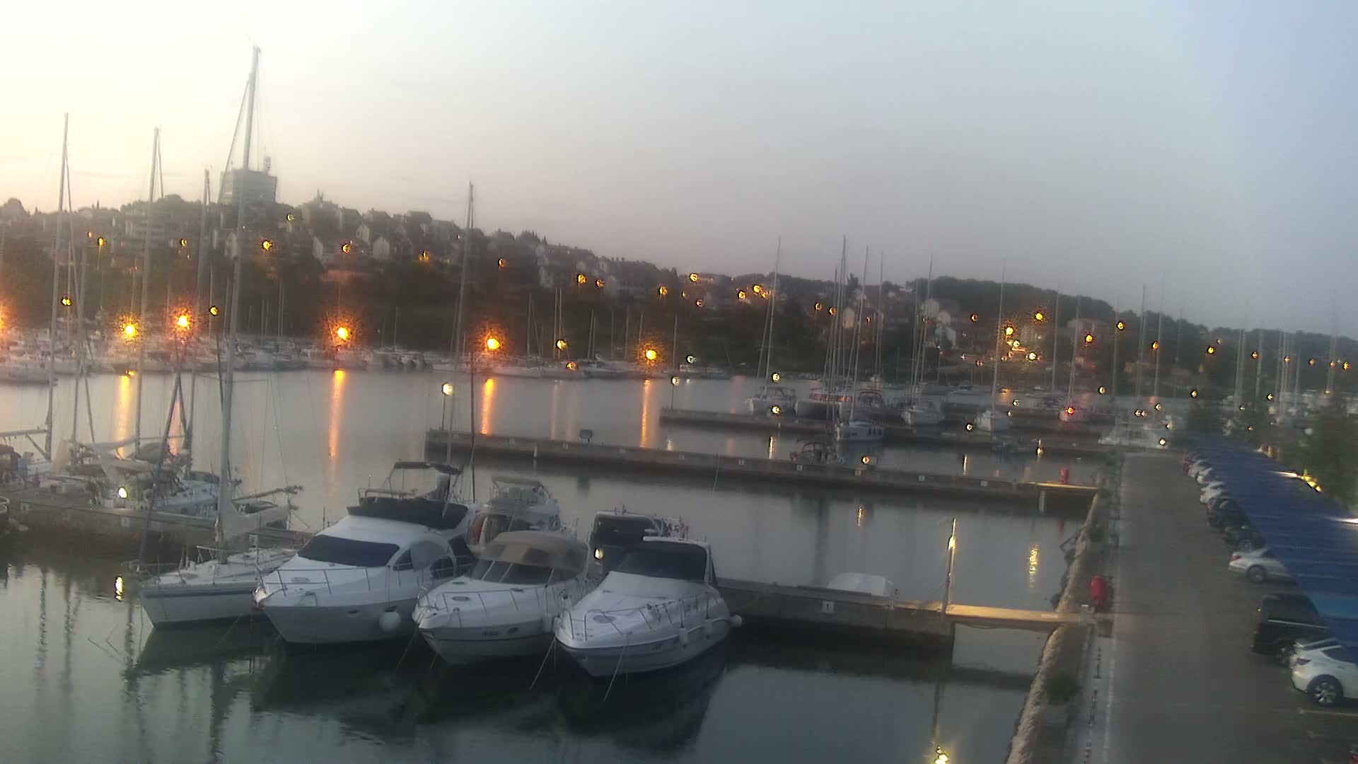 Several motorboats are docked at a marina at dusk, with many sailboats visible further in the harbor and a town illuminated with lights on a hill in the background.