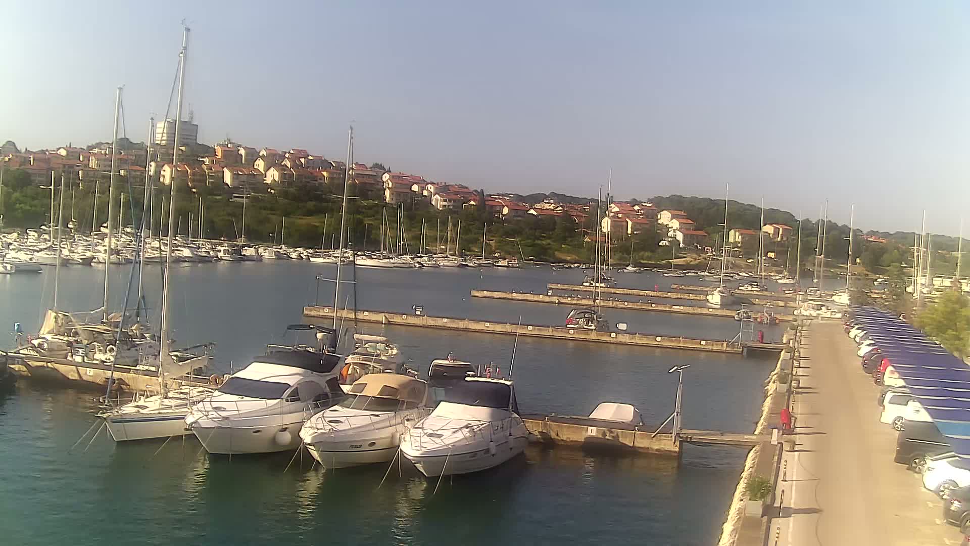 A marina filled with sailboats and motorboats is nestled next to a hillside town under a clear, sunny sky.