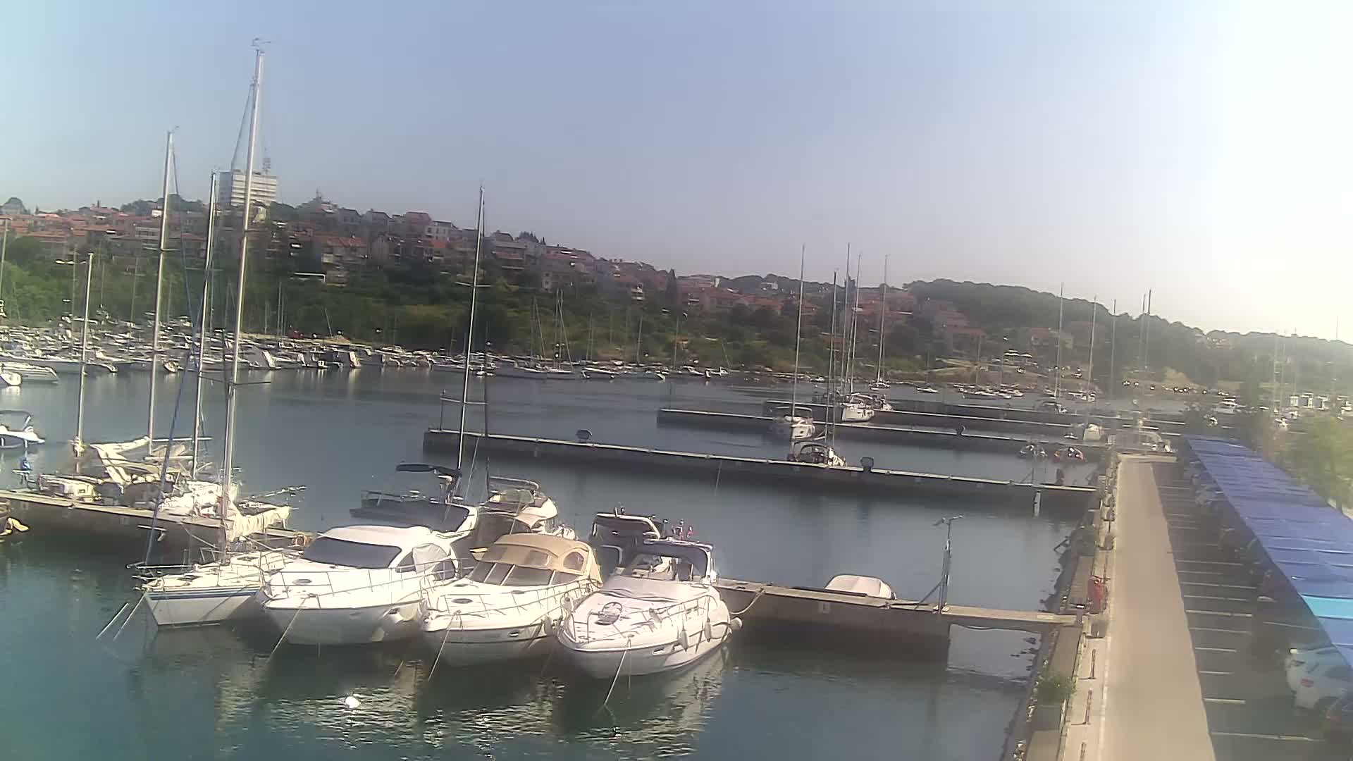 A marina filled with sailboats and motorboats on a calm, sunny day, with a town visible in the background.