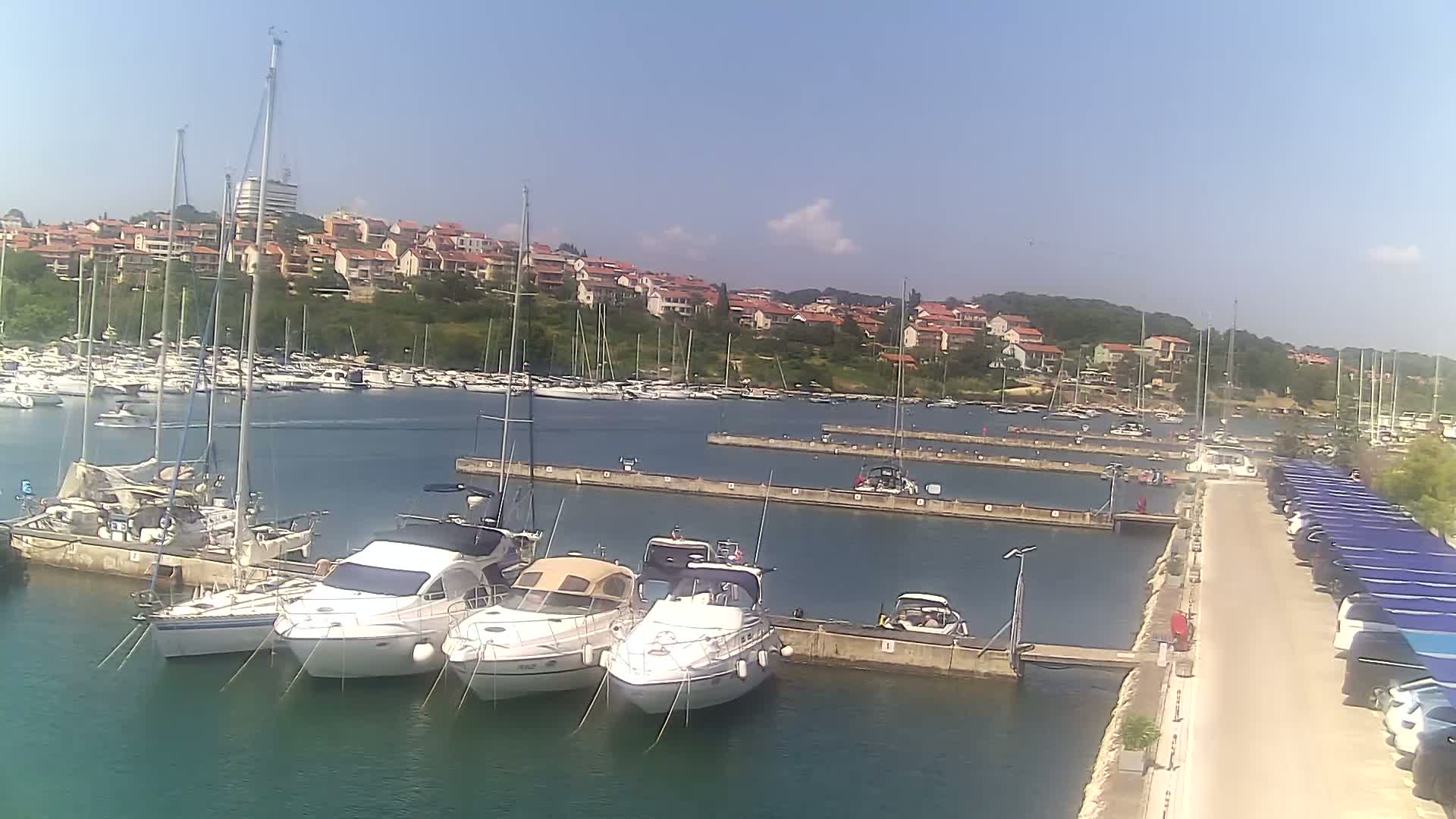 A marina filled with sailboats and motorboats is situated on calm water under a clear, sunny sky, with a hillside of houses visible in the background.