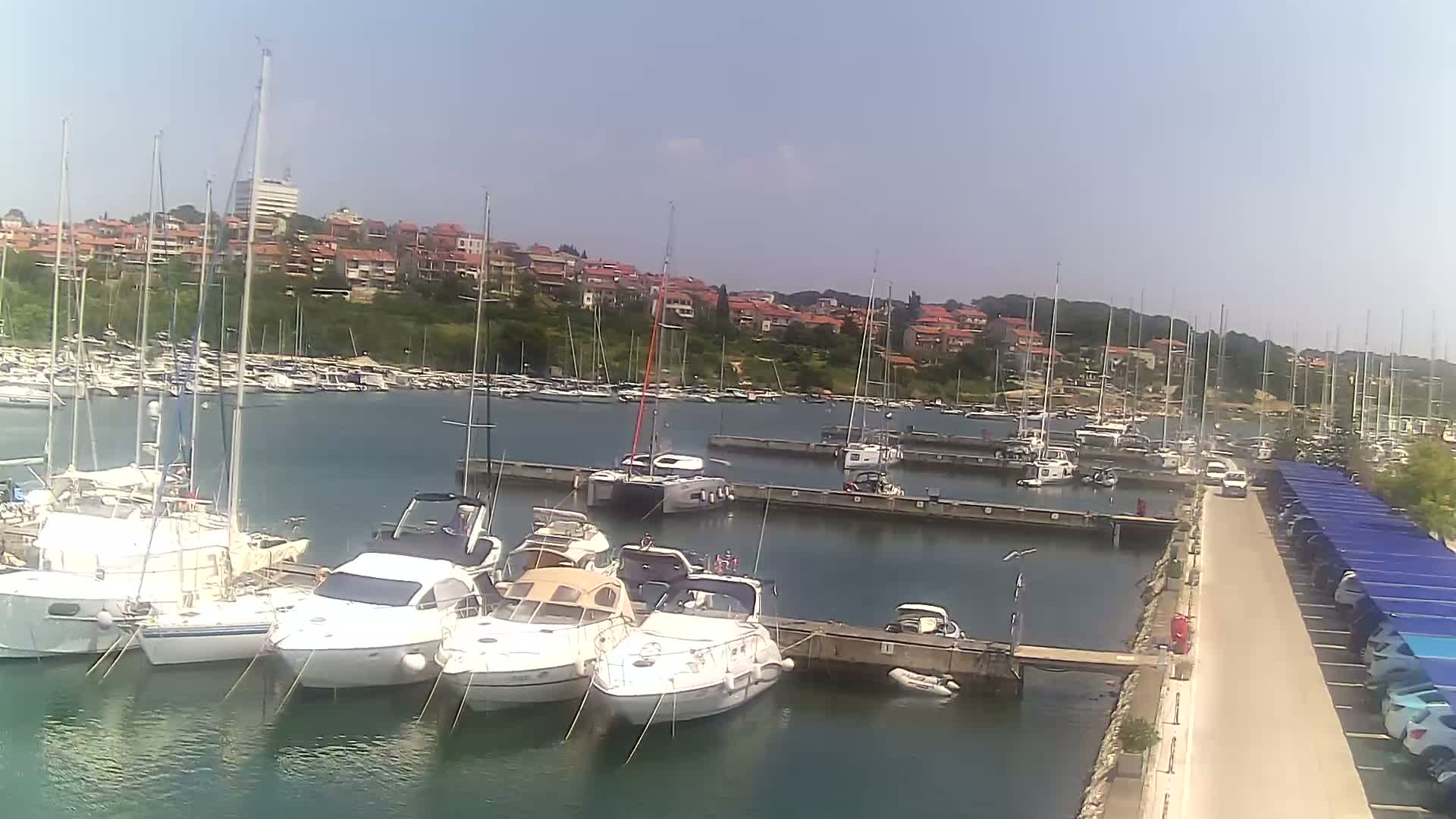 A marina filled with sailboats and motorboats on a sunny day, with a hillside town visible in the background.