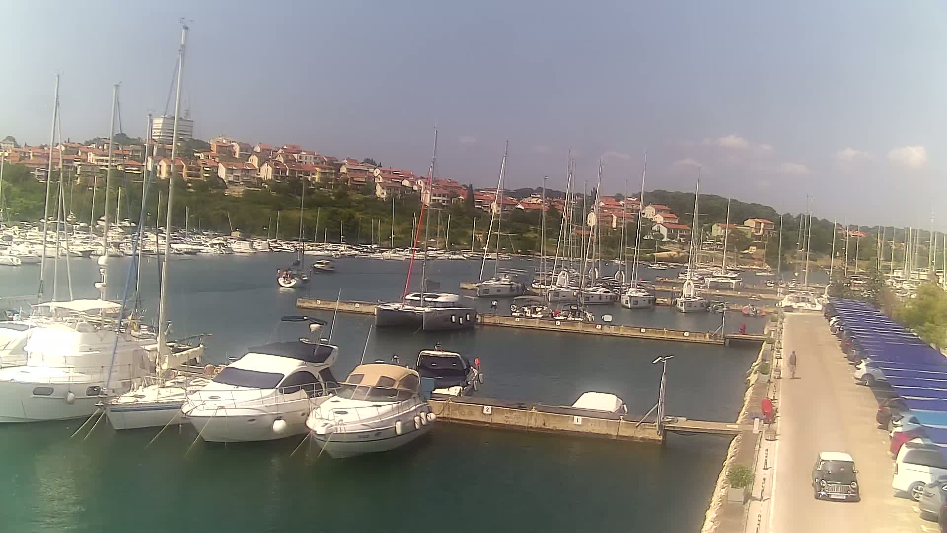 A marina filled with numerous sailboats and motorboats is situated next to a hillside town under a clear, sunny sky.