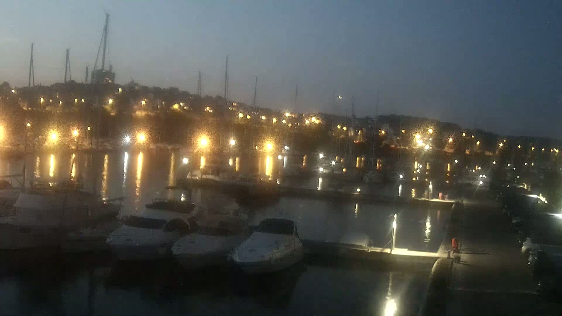 A marina at night, illuminated by many lights, with numerous boats moored alongside docks in calm water under a dark sky.