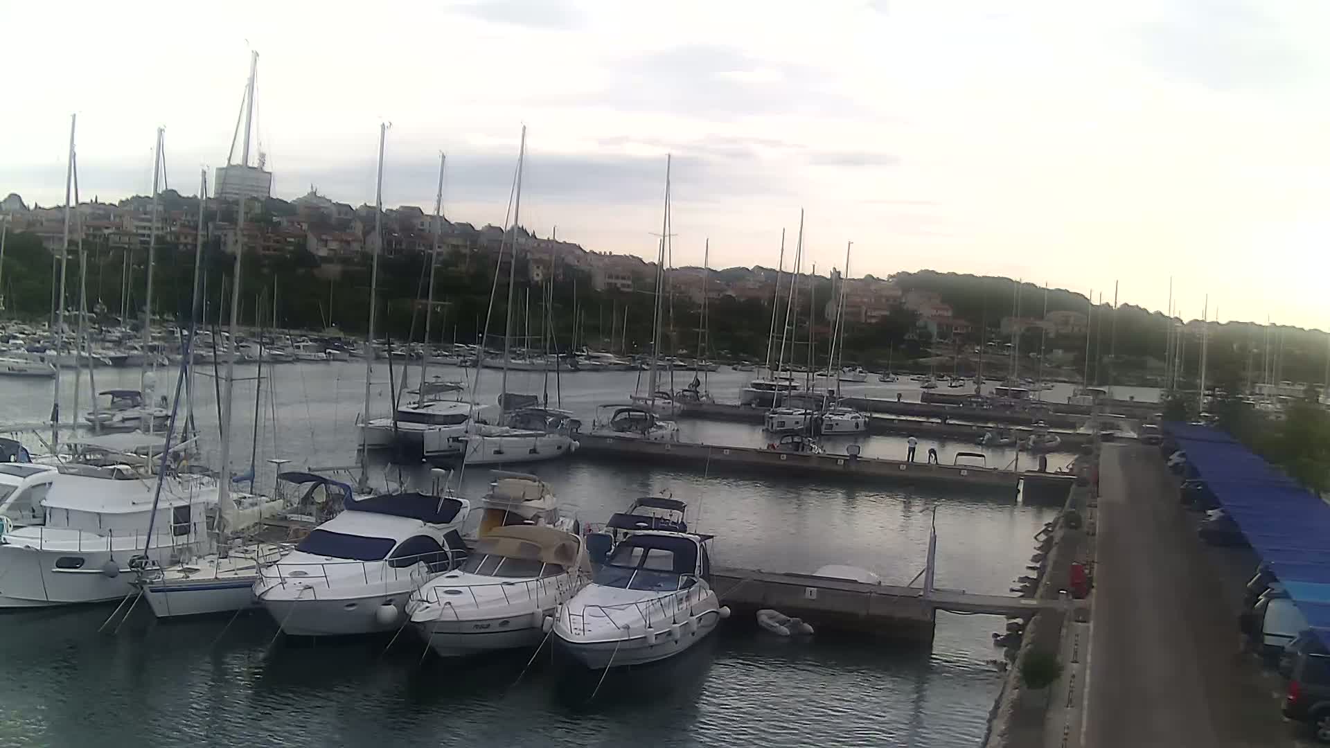 A marina filled with sailboats and motorboats is situated along a waterfront, under a partly cloudy sky.