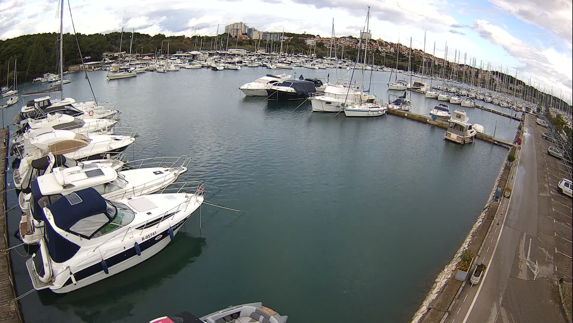 Numerous boats and yachts are moored in a bustling marina under a cloudy sky, with a waterfront road and parked cars visible on the right.