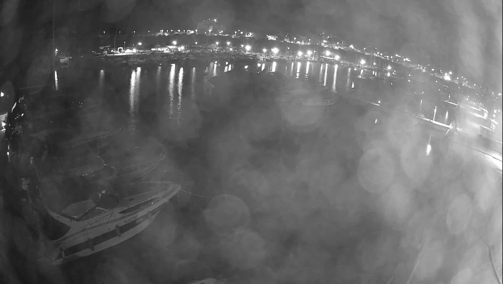 A blurry, grayscale, nighttime view reveals a marina or harbor filled with boats and a distant, lit shoreline reflecting on the water, observed through a camera lens obscured by rain.