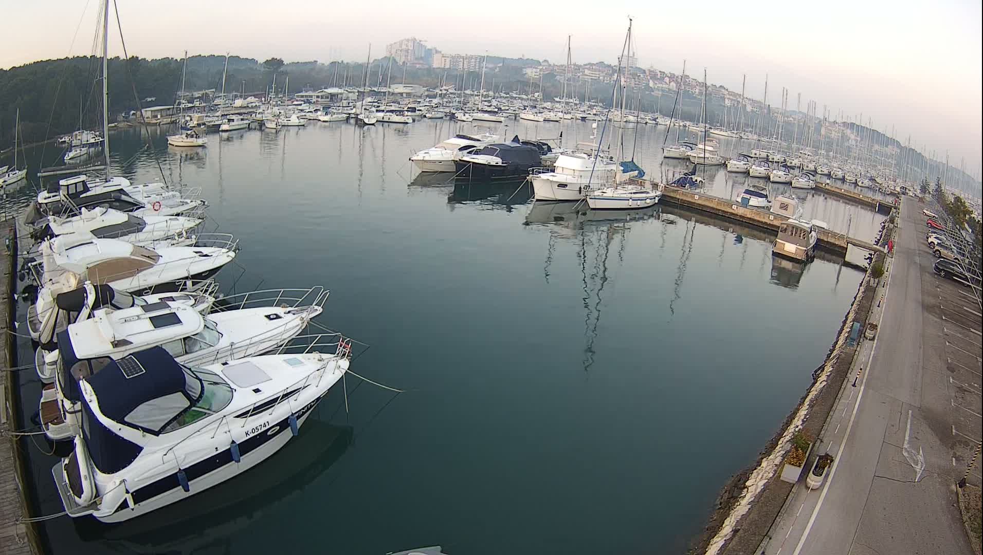 A blurry, grayscale, nighttime view reveals a marina or harbor filled with boats and a distant, lit shoreline reflecting on the water, observed through a camera lens obscured by rain.
