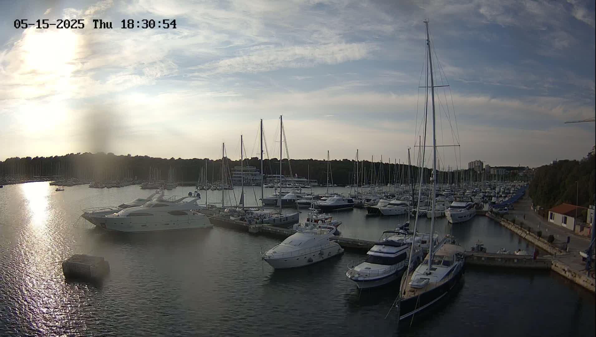 A marina filled with numerous sailboats and motor yachts is seen at sunset under partly cloudy skies.
