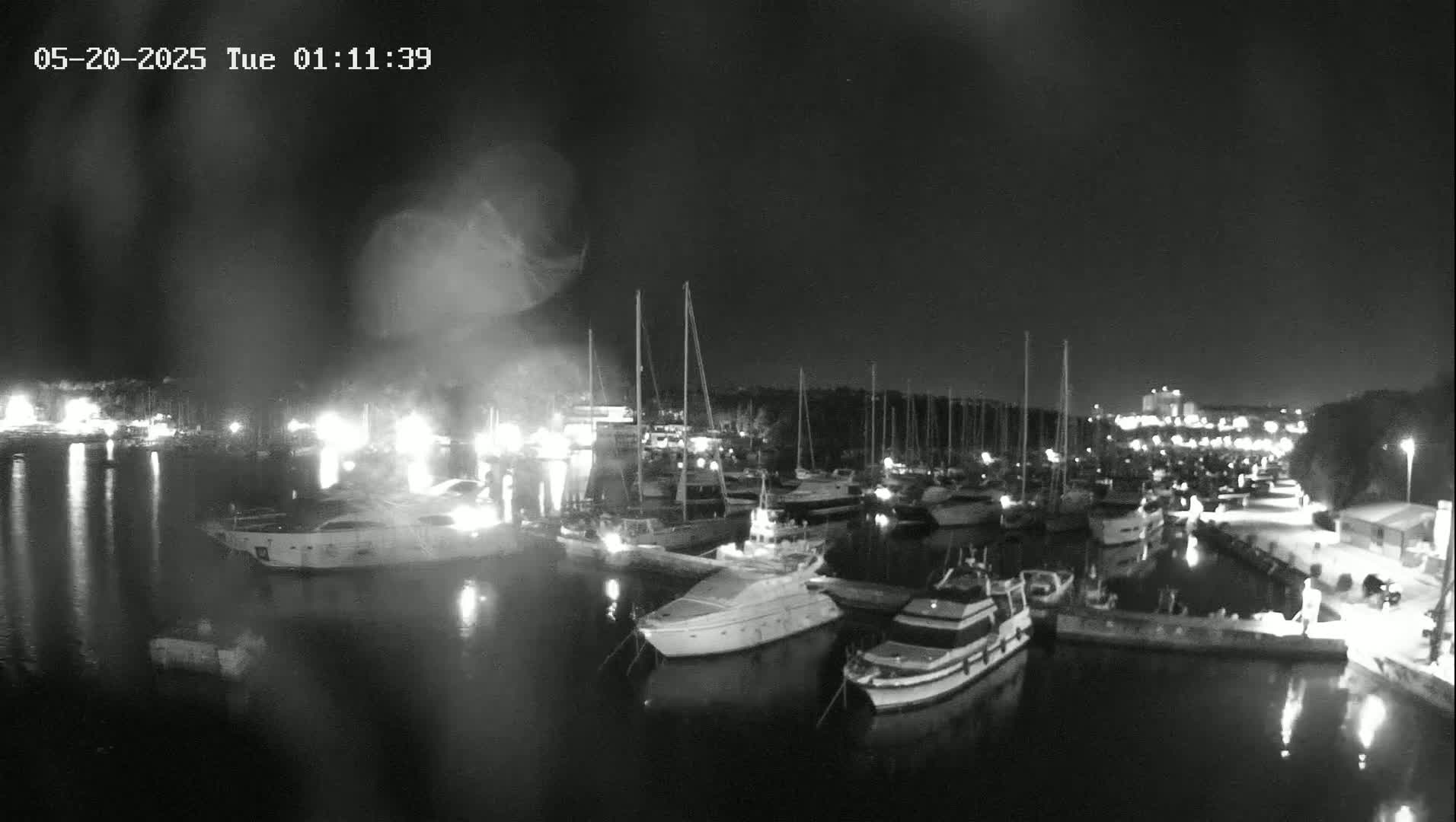 A nighttime, rain-streaked view of a marina filled with numerous yachts and sailboats docked along a waterfront.