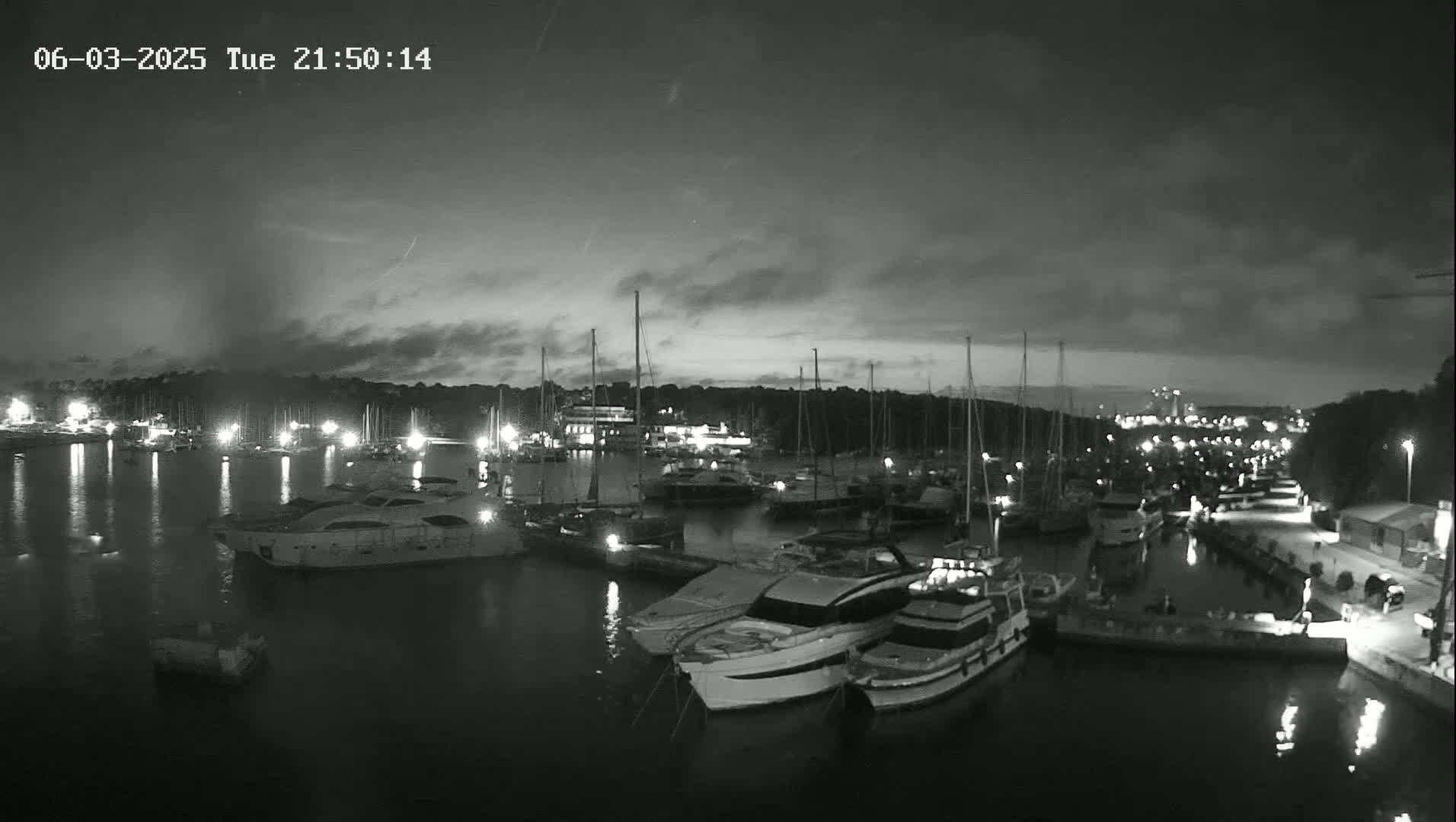 A nighttime, overcast view of a marina filled with docked sailboats and motorboats, illuminated by various lights.