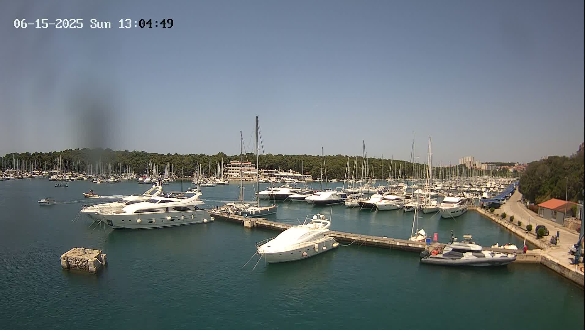 A sunny day at a marina shows numerous yachts and sailboats docked, with several larger motor yachts prominent in the foreground.