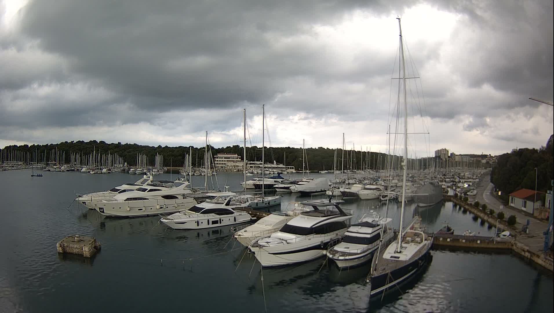A crowded marina, filled with numerous motor yachts and sailboats, is seen under a heavily overcast and gloomy sky, with a lush forested coastline and distant buildings framing the harbor.