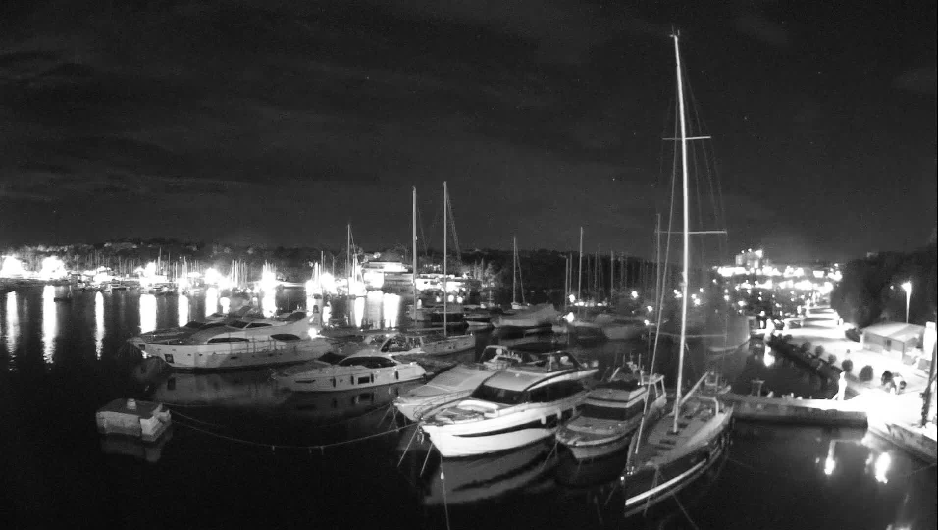 A black and white night view displays a marina densely packed with various boats and yachts, their lights reflecting on the calm water beneath a cloudy sky.