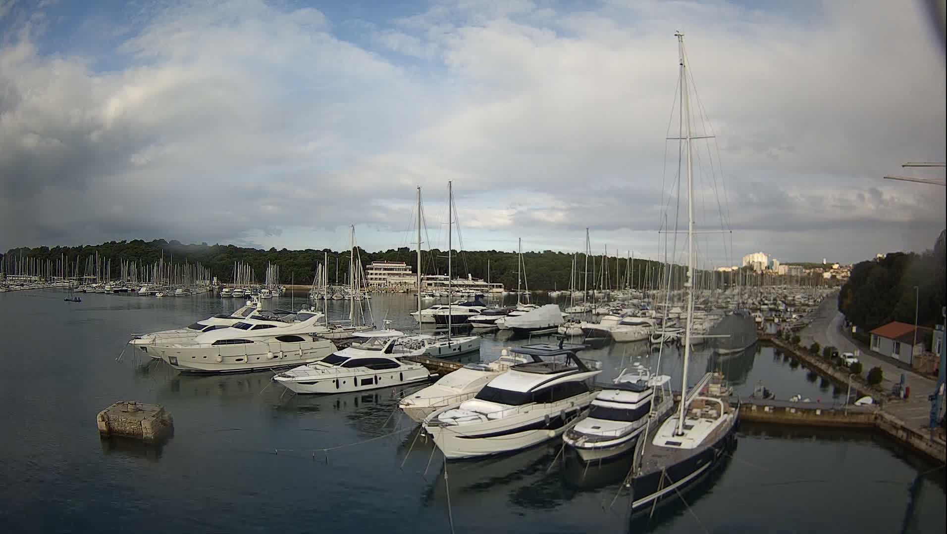 Numerous sailboats and motor yachts are docked in a bustling marina, flanked by a tree-lined shore and a distant town, under an overcast sky with patches of blue.