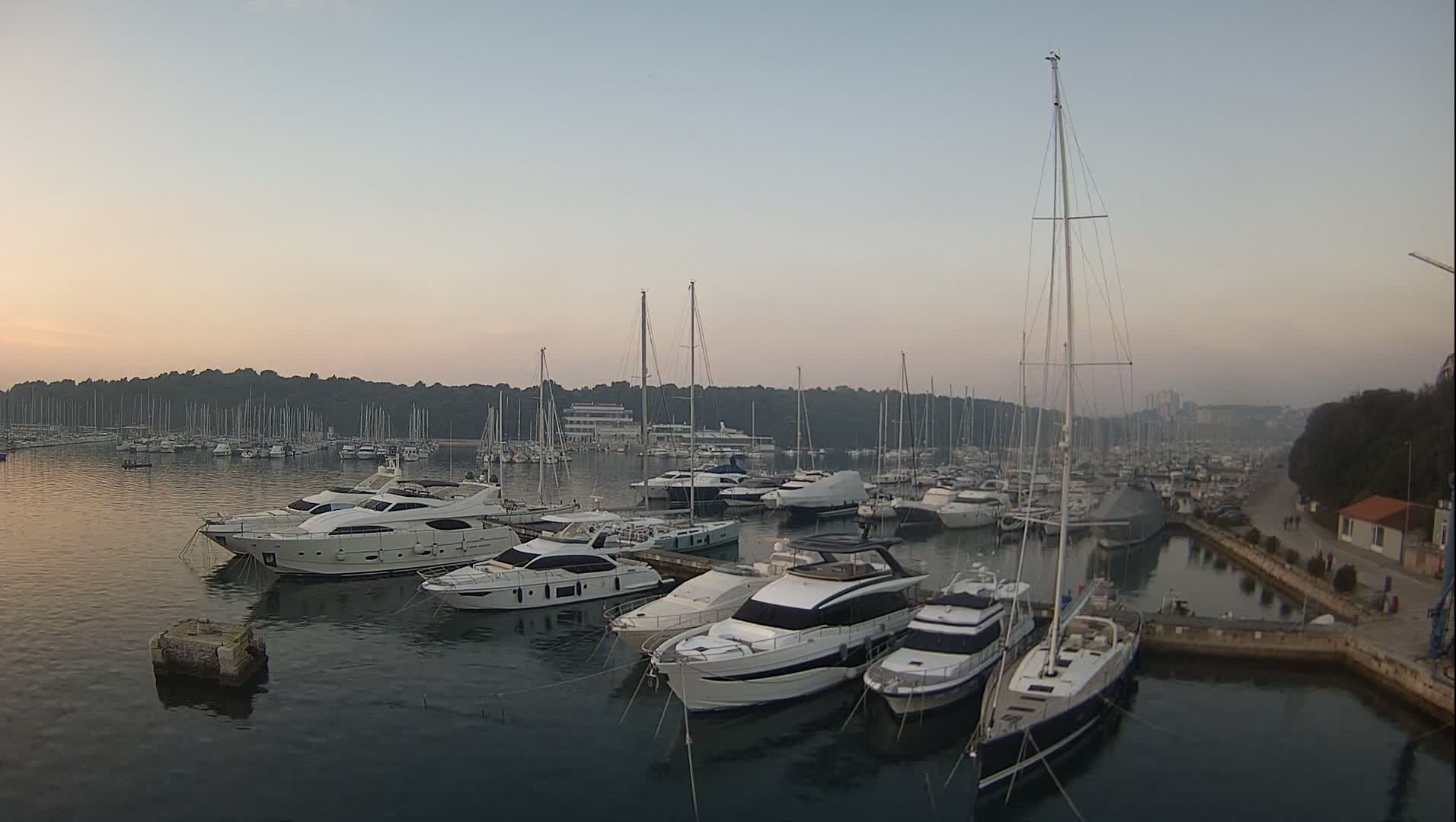Numerous sailboats and motor yachts are docked in a bustling marina, flanked by a tree-lined shore and a distant town, under an overcast sky with patches of blue.