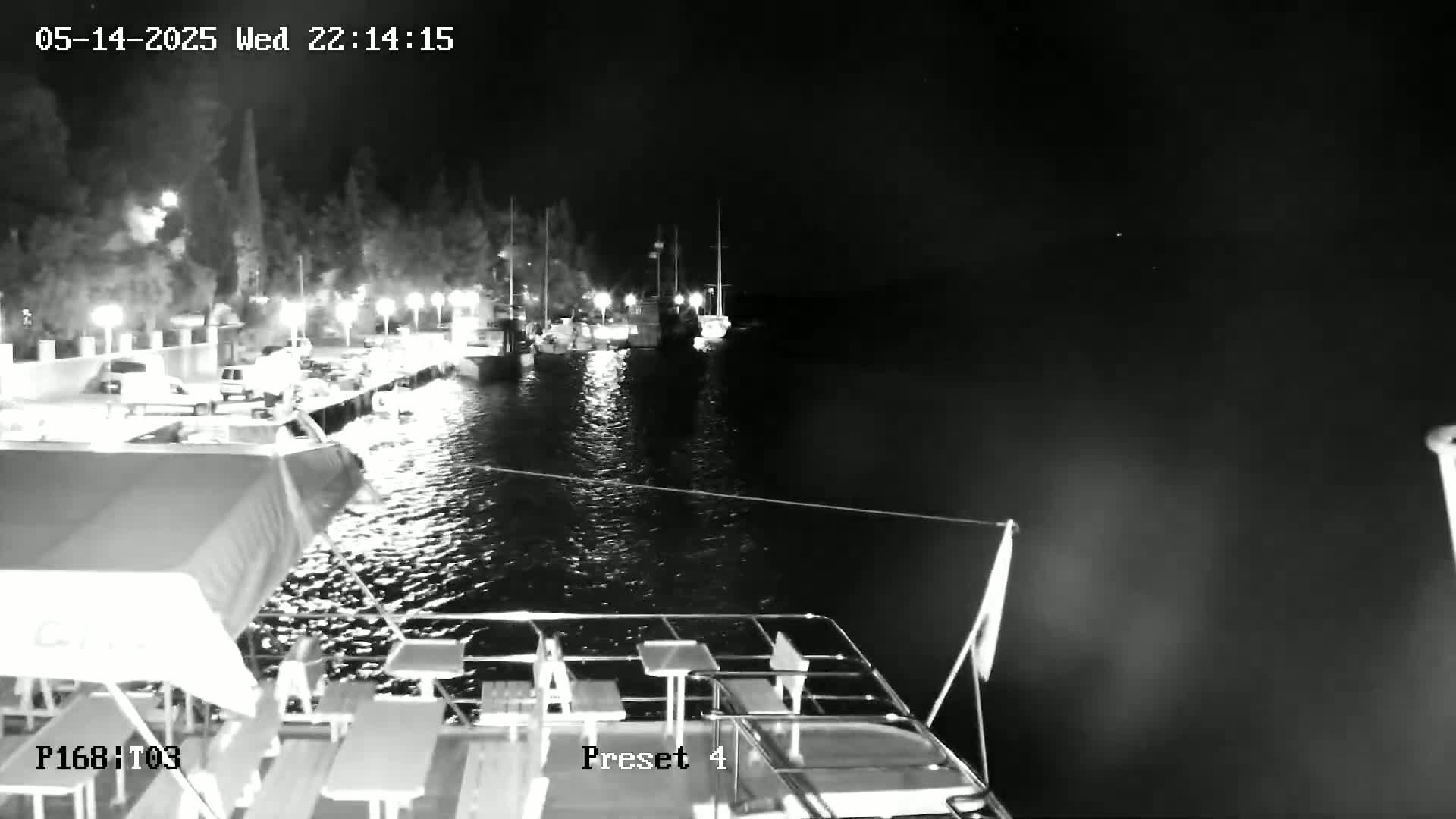 A nighttime view from a boat shows a calm body of water with several boats docked along a shore illuminated by lights.