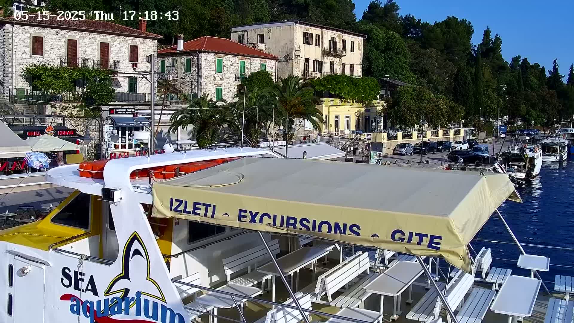A boat with a beige awning and white benches sits in a harbor next to buildings and parked cars on a sunny day.