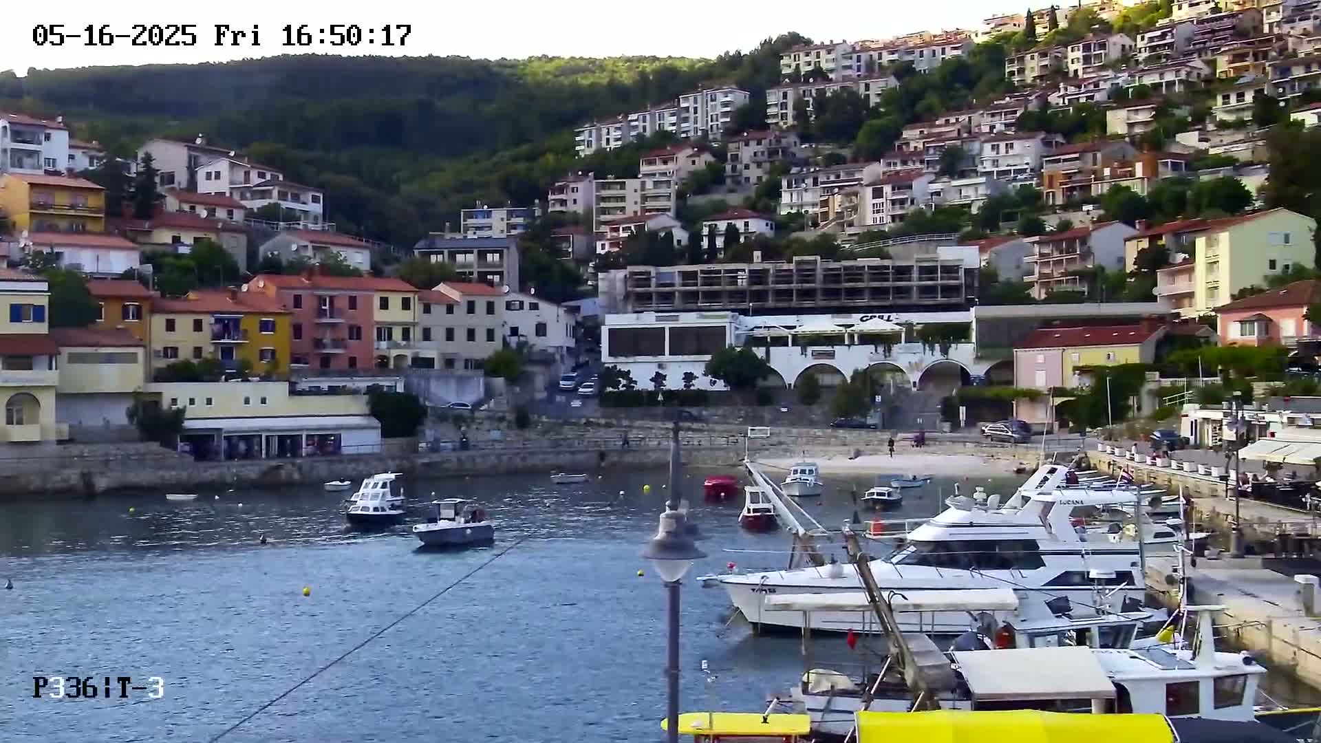 A small harbor filled with boats is nestled beside a hillside town of multi-story buildings under a partly cloudy sky.