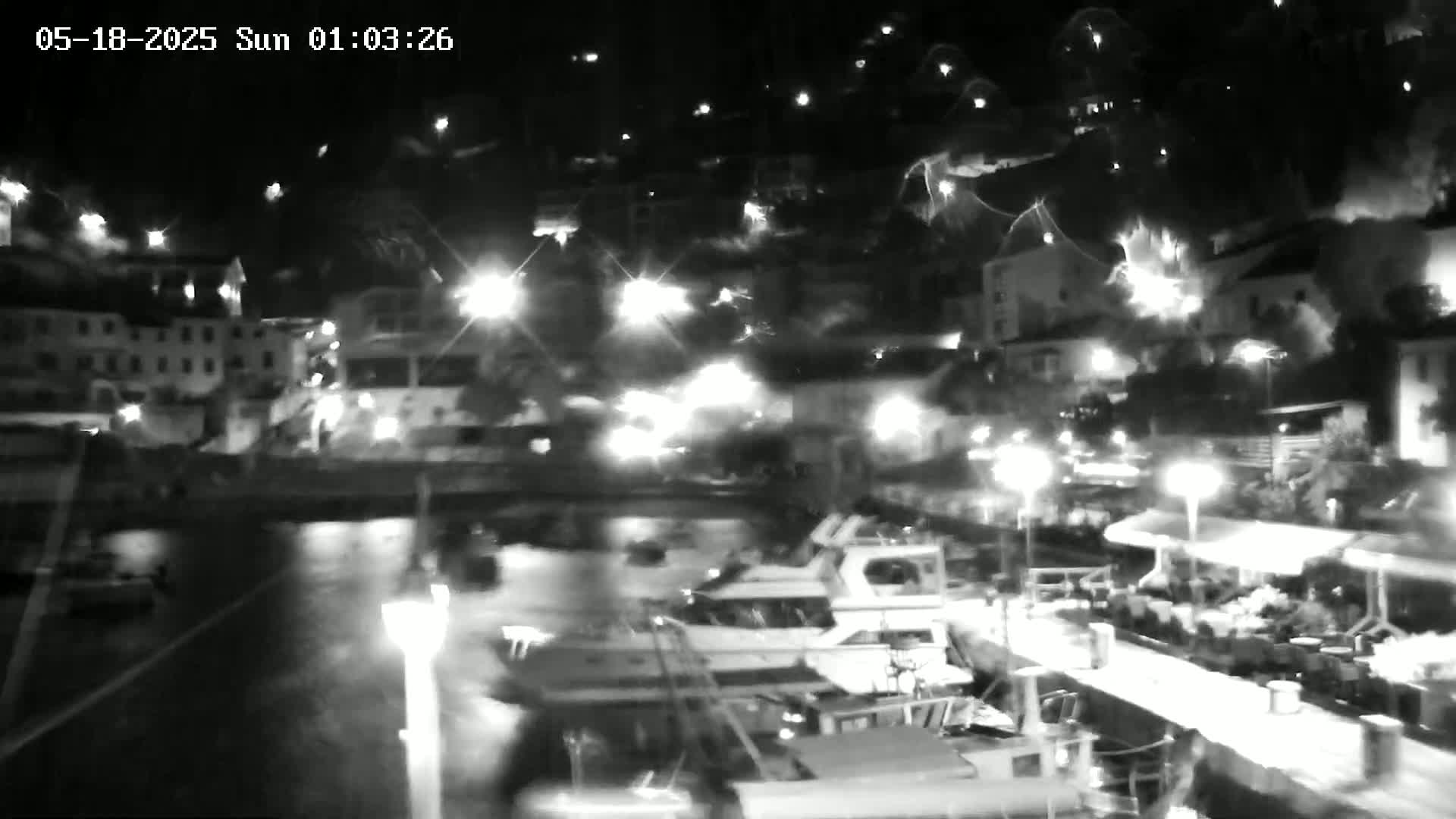 A nighttime, low-light view of a harbor filled with boats, alongside a waterfront with buildings and outdoor seating areas illuminated by streetlights.