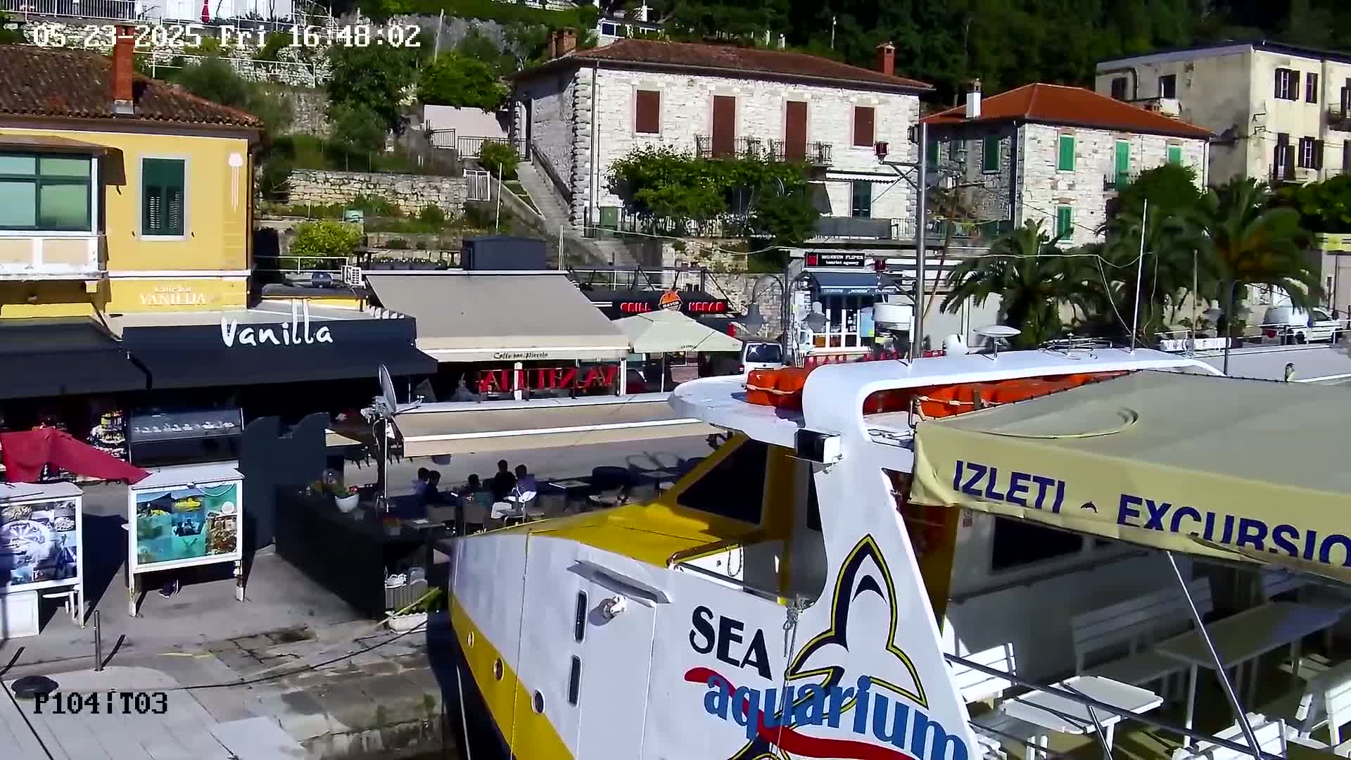 A yellow and white excursion boat is docked near a waterfront area with stone buildings, cafes, and palm trees on a sunny day.