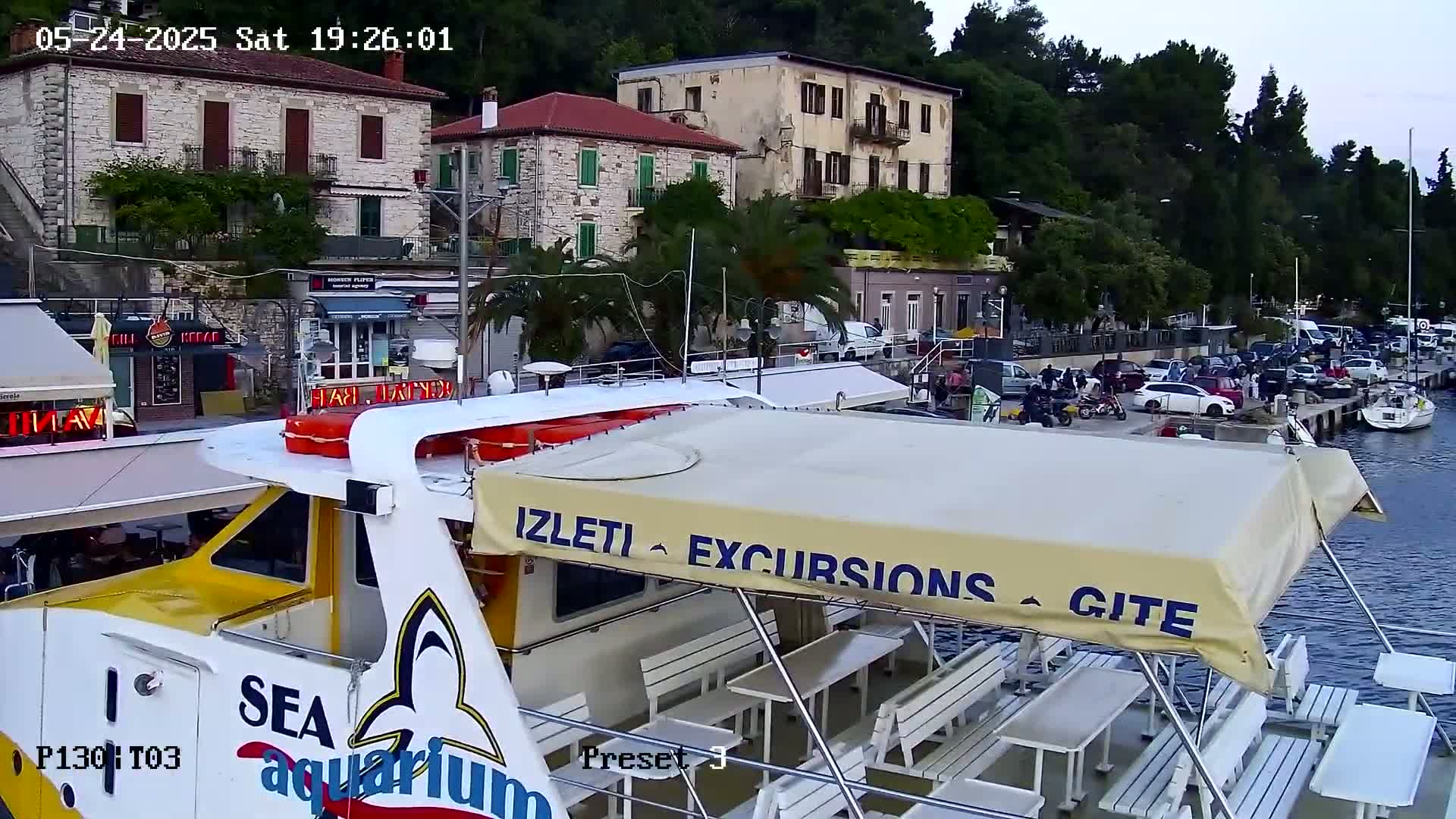 A yellow and white excursion boat with white tables and benches is docked near several buildings and parked cars, under an overcast sky.