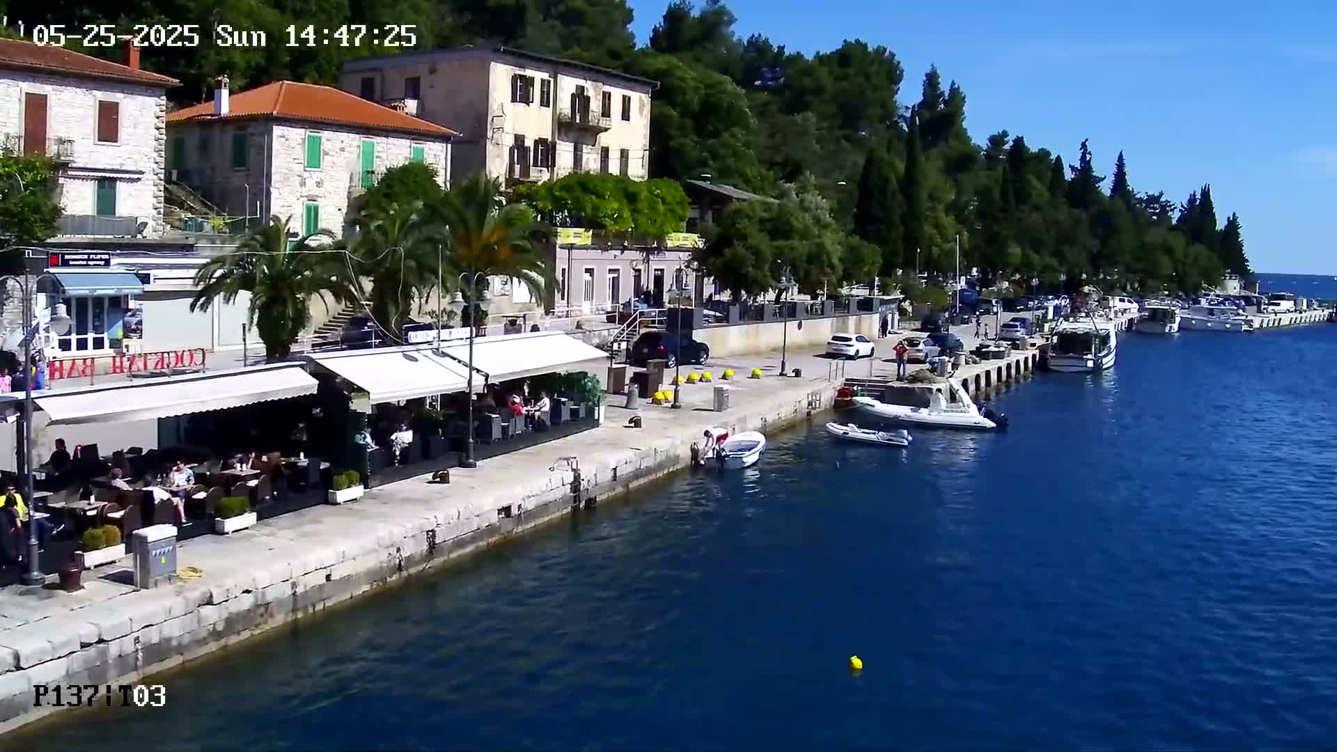 A waterfront view on a sunny day shows a stone building, several outdoor cafes, parked cars, and numerous boats moored along a pier.