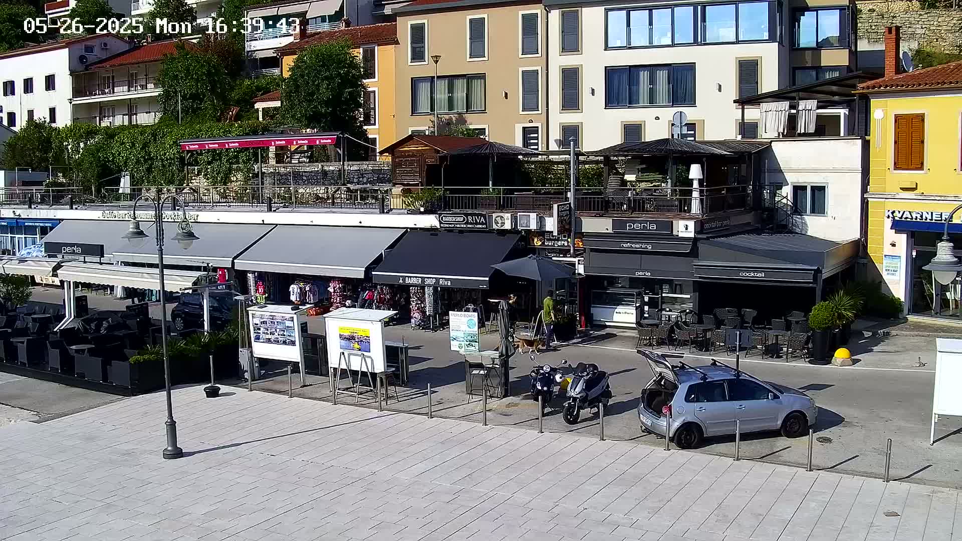 A sunny day overlooking a paved waterfront area with several shops and restaurants under awnings, a few parked vehicles, and motorcycles.