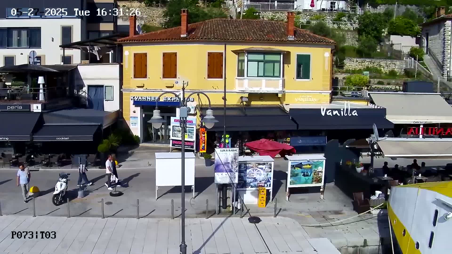 A sunny outdoor scene shows a paved waterfront area with several shops and restaurants, some people walking by, and a parked scooter.