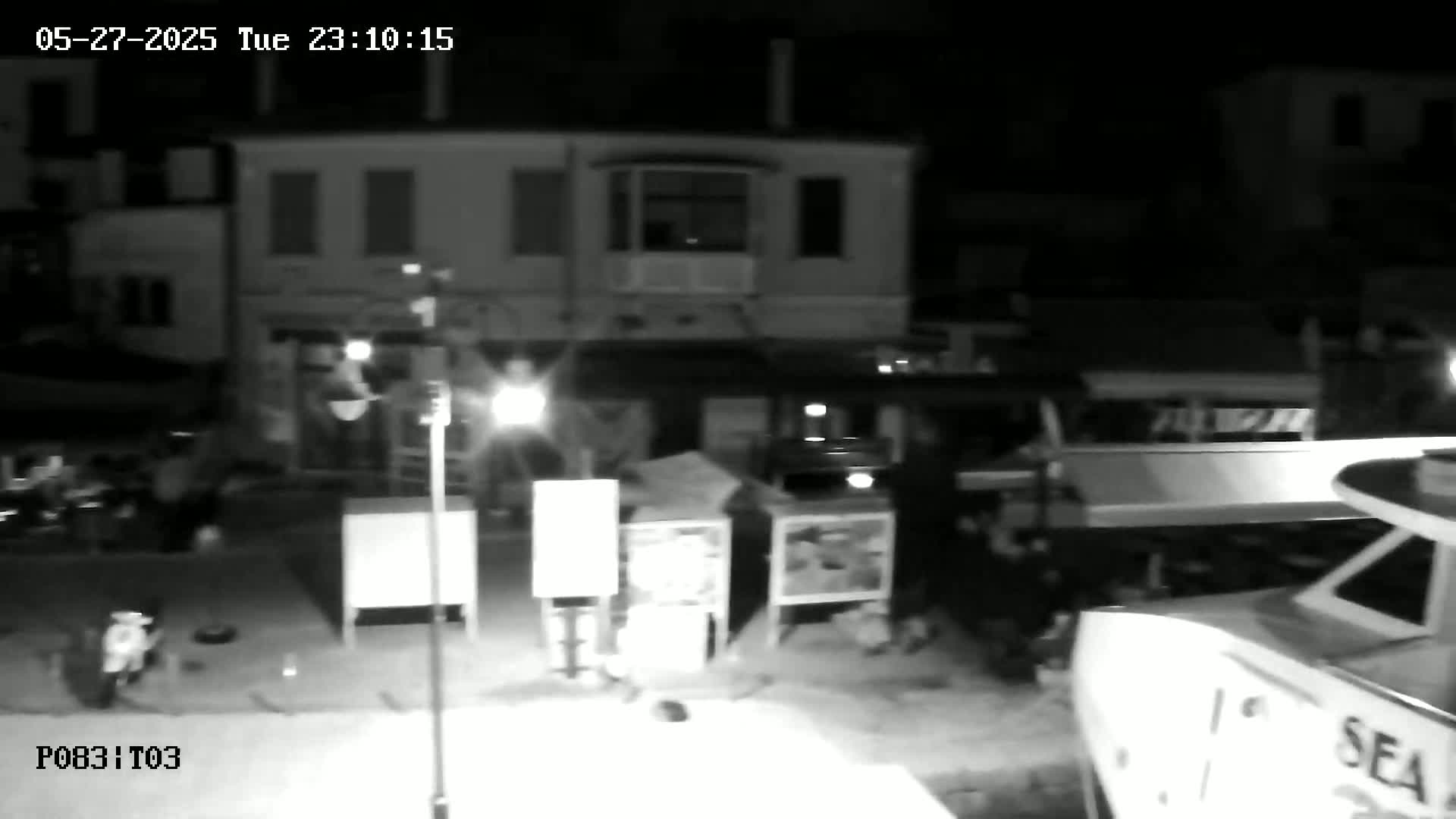 A nighttime, black-and-white security camera view shows a street scene with several people near buildings and boats.