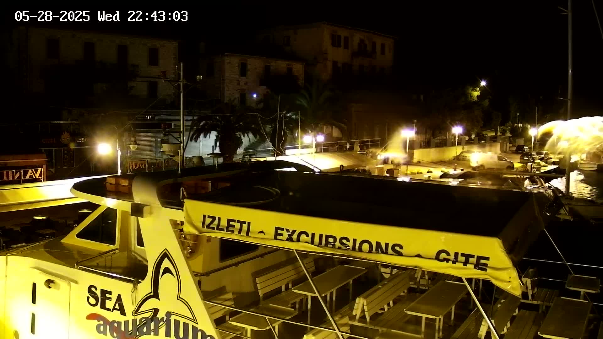 A yellow excursion boat is docked at night near buildings with palm trees, under partly cloudy skies, with water splashing nearby.