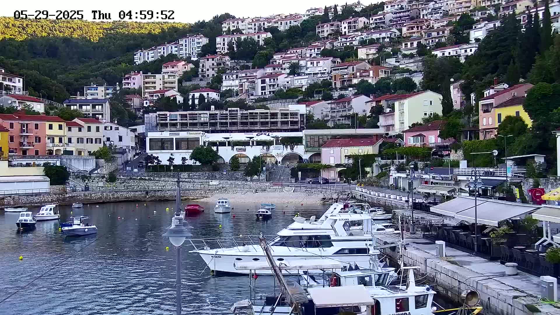 A small harbor filled with boats is nestled below a hillside densely packed with multi-story buildings, on a partly cloudy day.