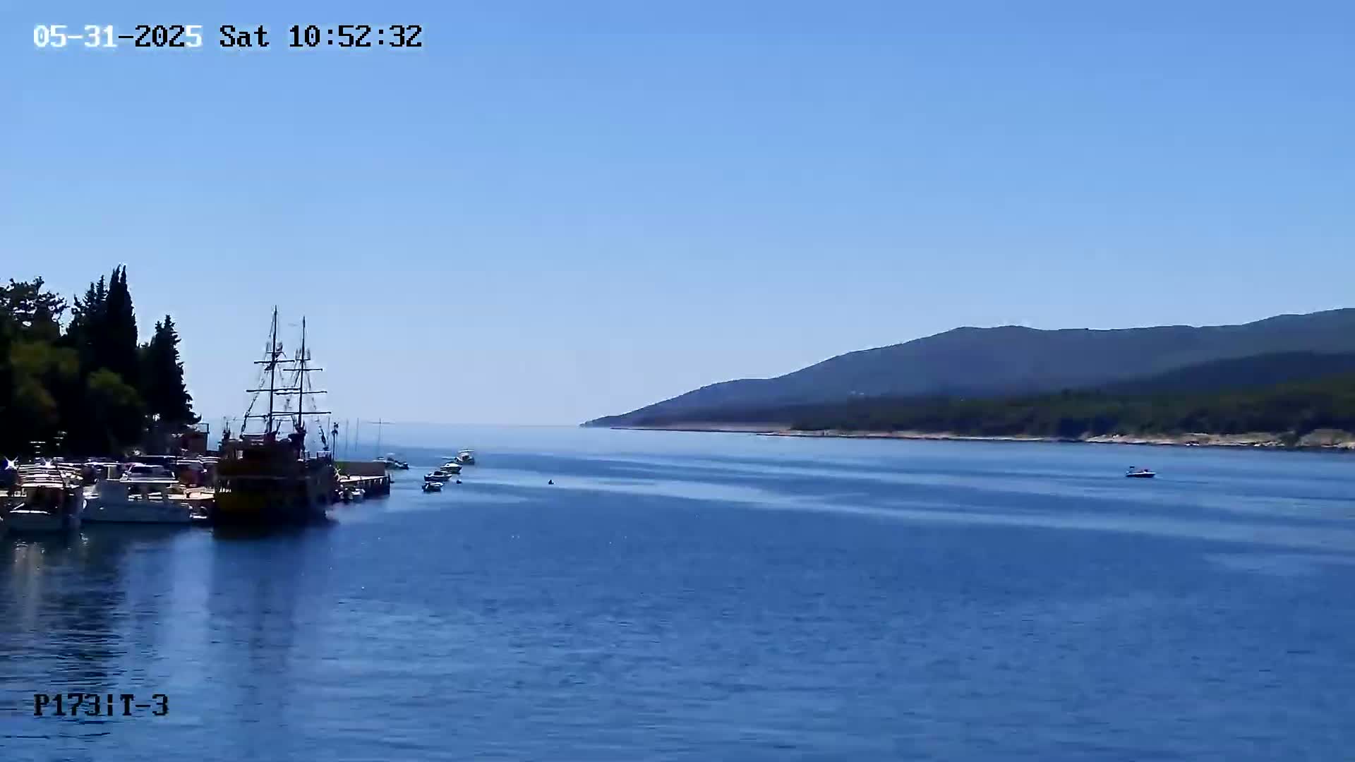 Several boats are moored near a shoreline with dark green trees, under a clear blue sky, with mountains visible in the distance.