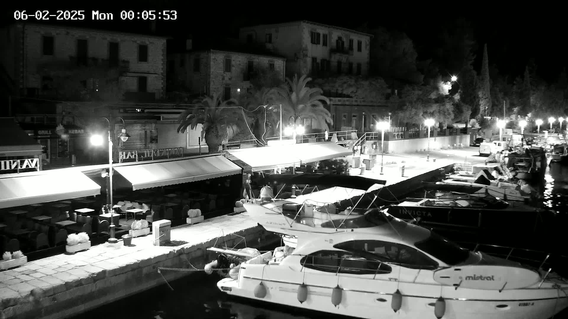 A nighttime, black-and-white view of a harbor with docked boats, outdoor seating areas at waterfront restaurants, and buildings lit by streetlights.