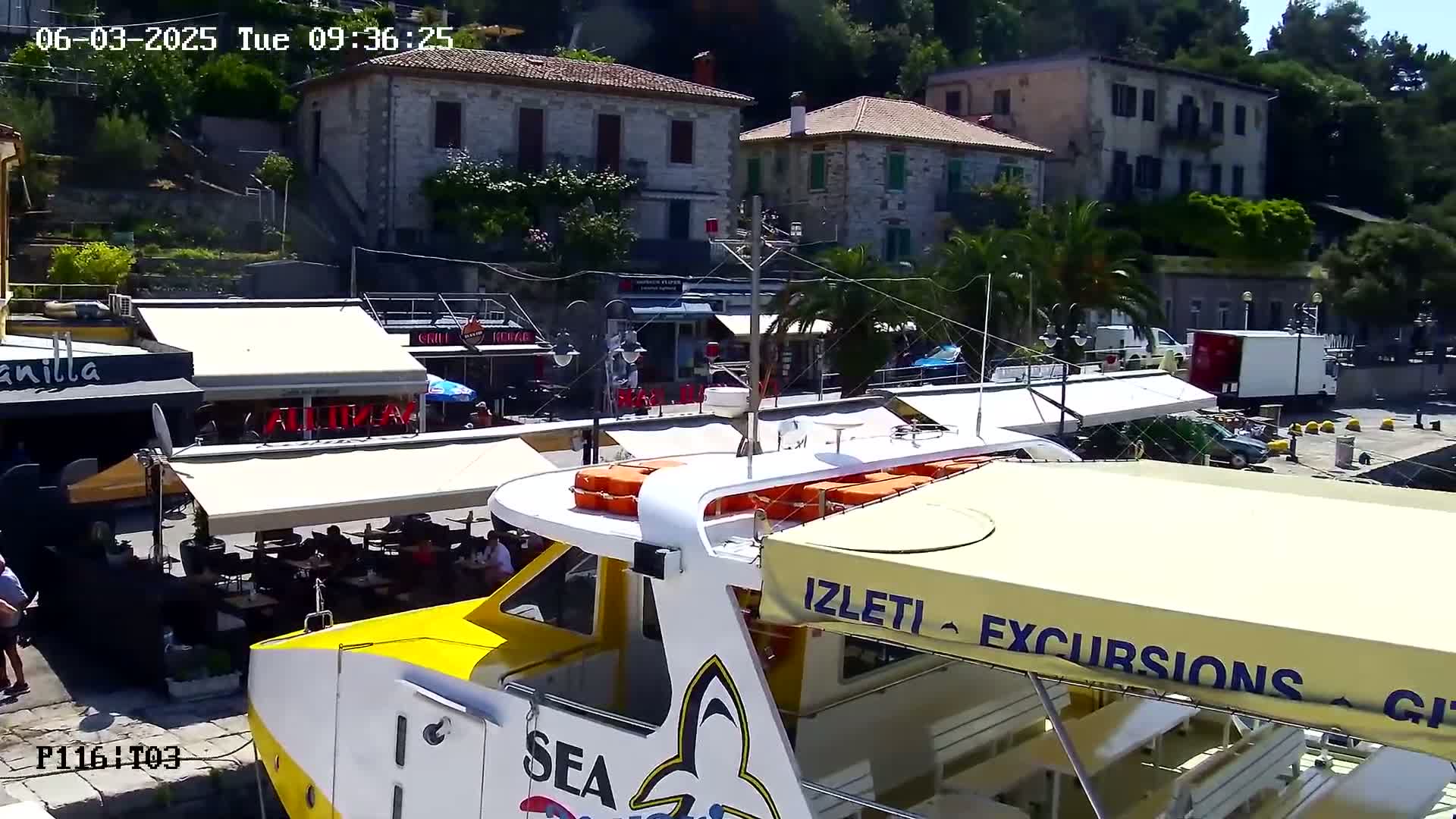 A yellow and white excursion boat is docked in a harbor next to stone buildings and outdoor cafes under a sunny sky.