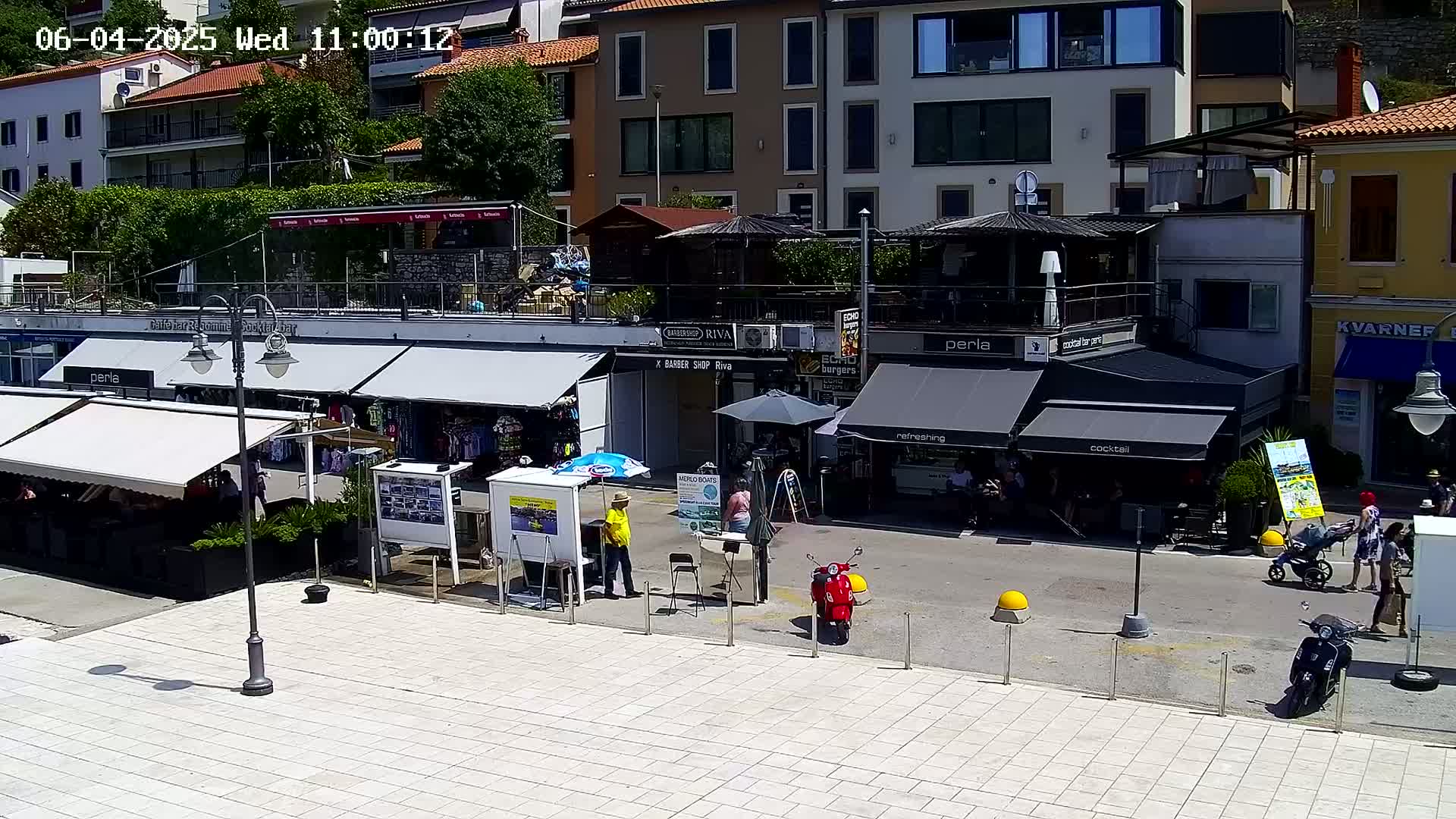 A sunny outdoor plaza scene with several shops and restaurants, some people walking around, and a red scooter parked.