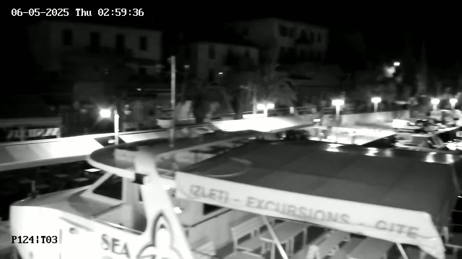 A nighttime, low-angle view shows the top of a boat with several people walking on a dock nearby.
