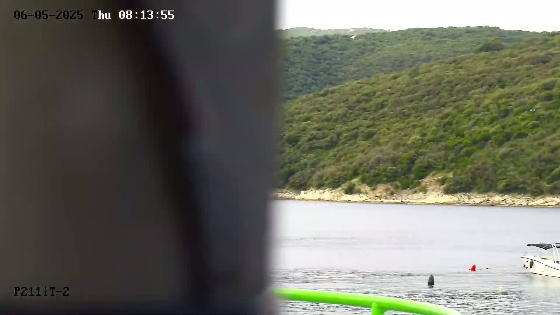 A partially obscured view shows a calm body of water with a small boat and a green railing in the foreground, next to a rocky, green hillside.