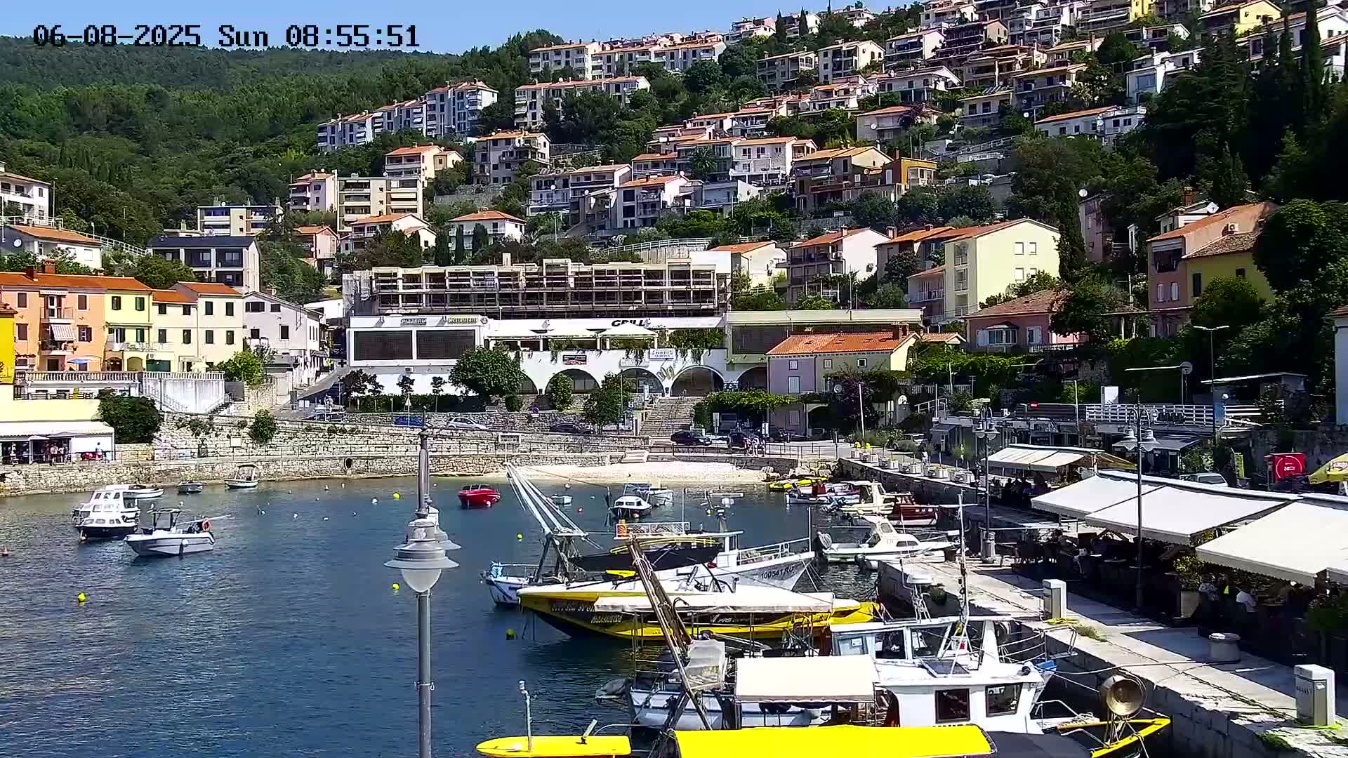 A harbor filled with small boats is nestled between a hillside of buildings and a waterfront promenade with outdoor seating under awnings on a sunny day.