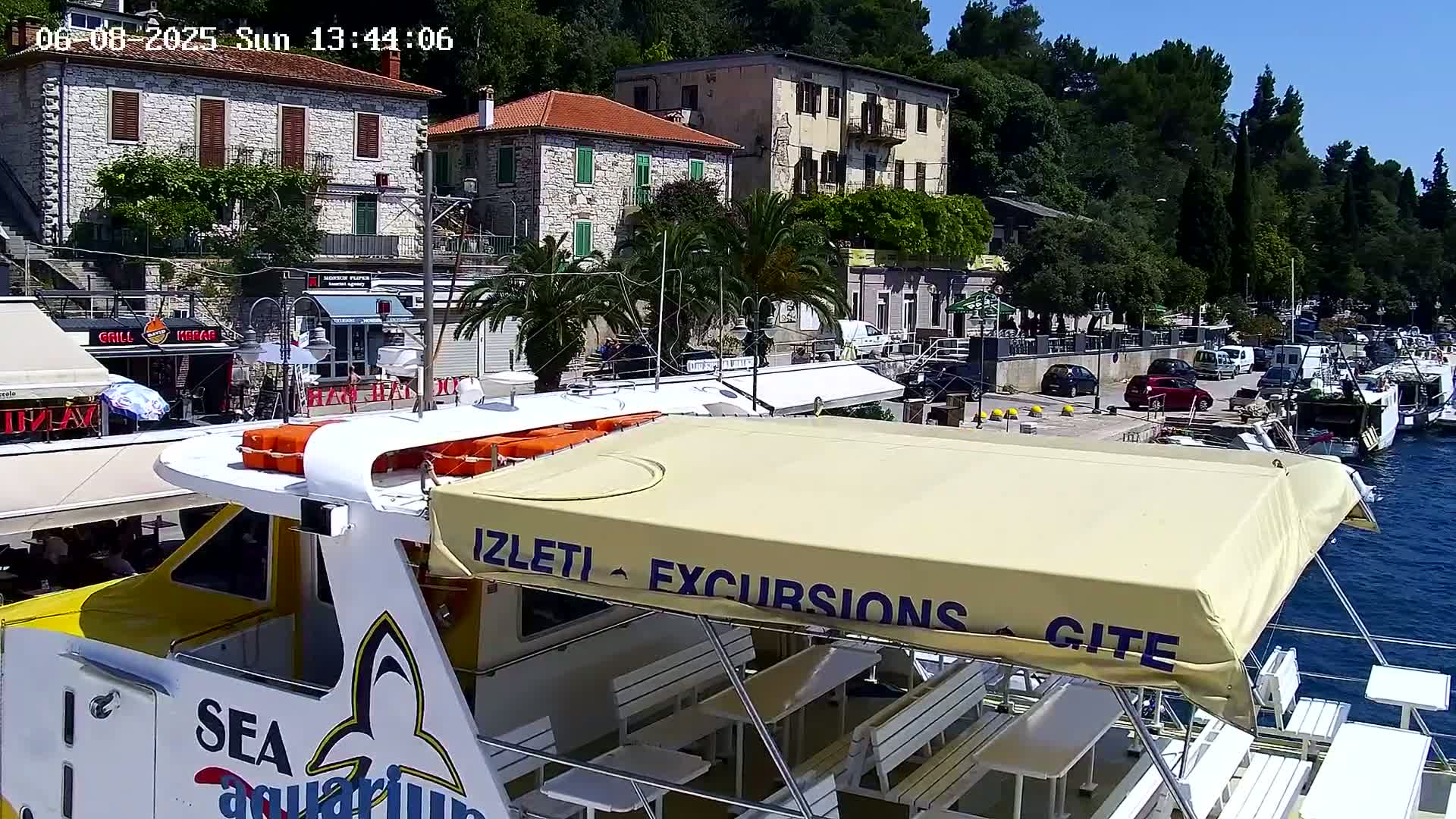 A yellow and white passenger boat with a tan awning is docked at a harbor, overlooking buildings and parked cars on a sunny day.