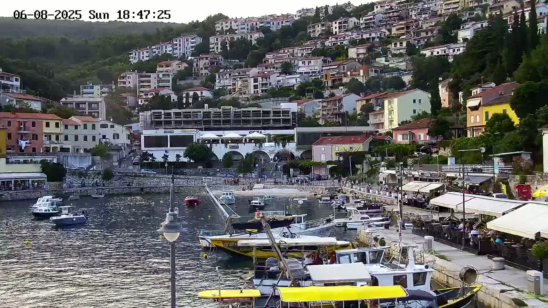 A harbor filled with boats is nestled below a hillside town of colorful buildings under a partly cloudy sky.